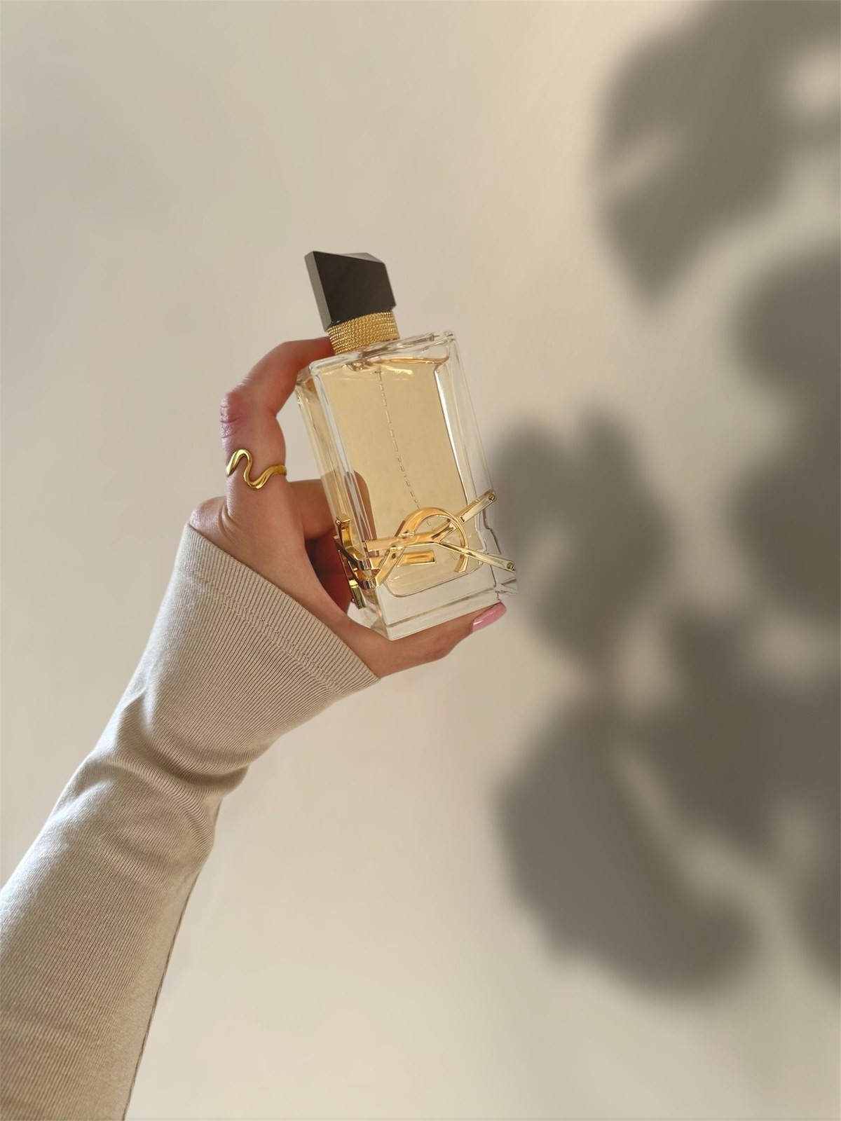 Hand in a cream sweater holding up a YSL Libre perfume bottle with a floral shadow behind it