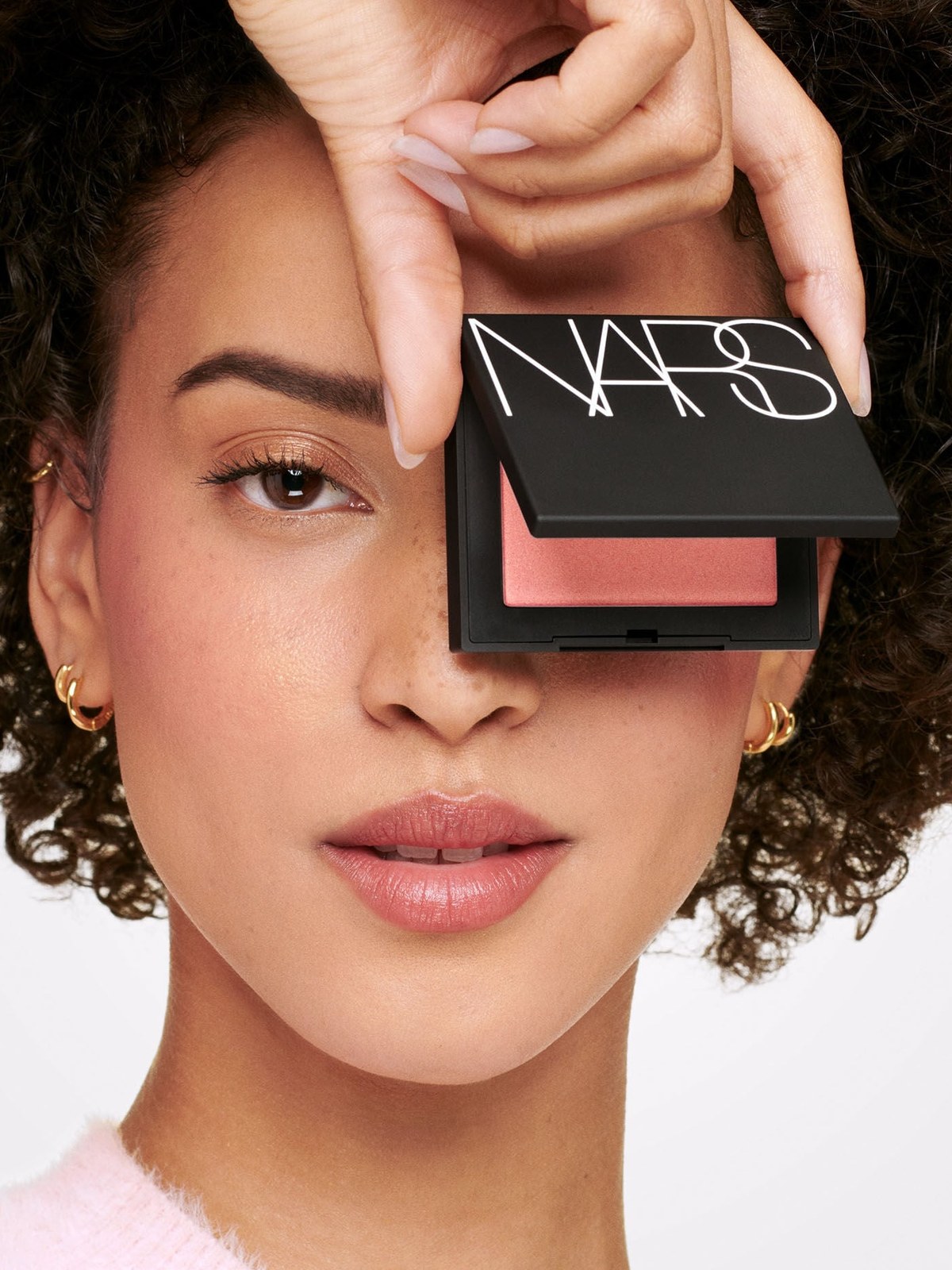 Curly haired model holding a NARS blush compact over one eye