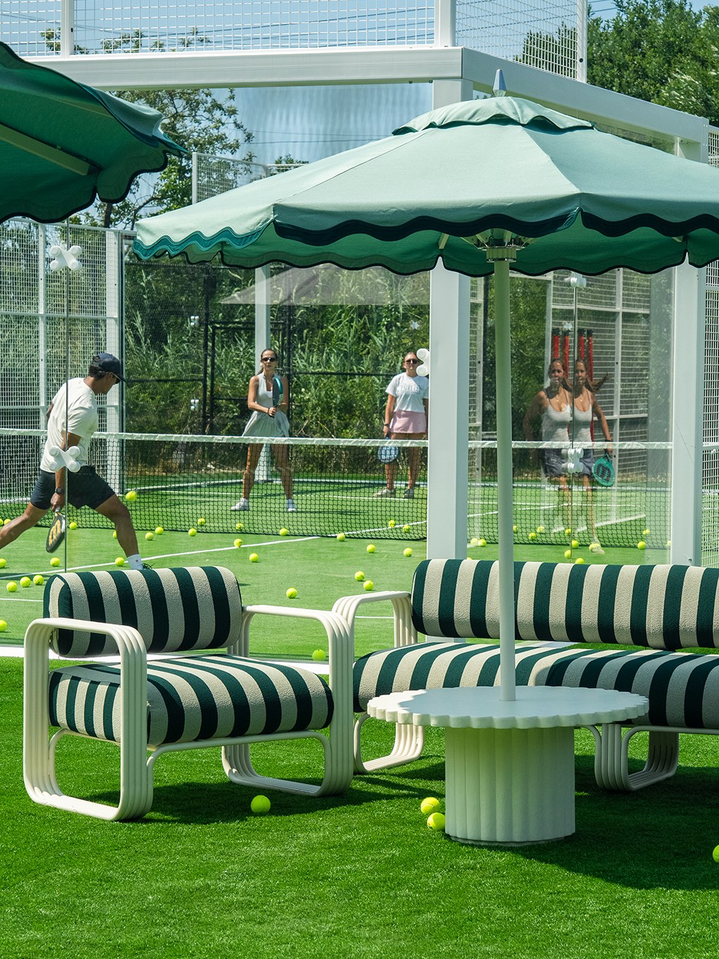Green striped outdoor seating beneath a scalloped umbrella while adults play tennis in the background