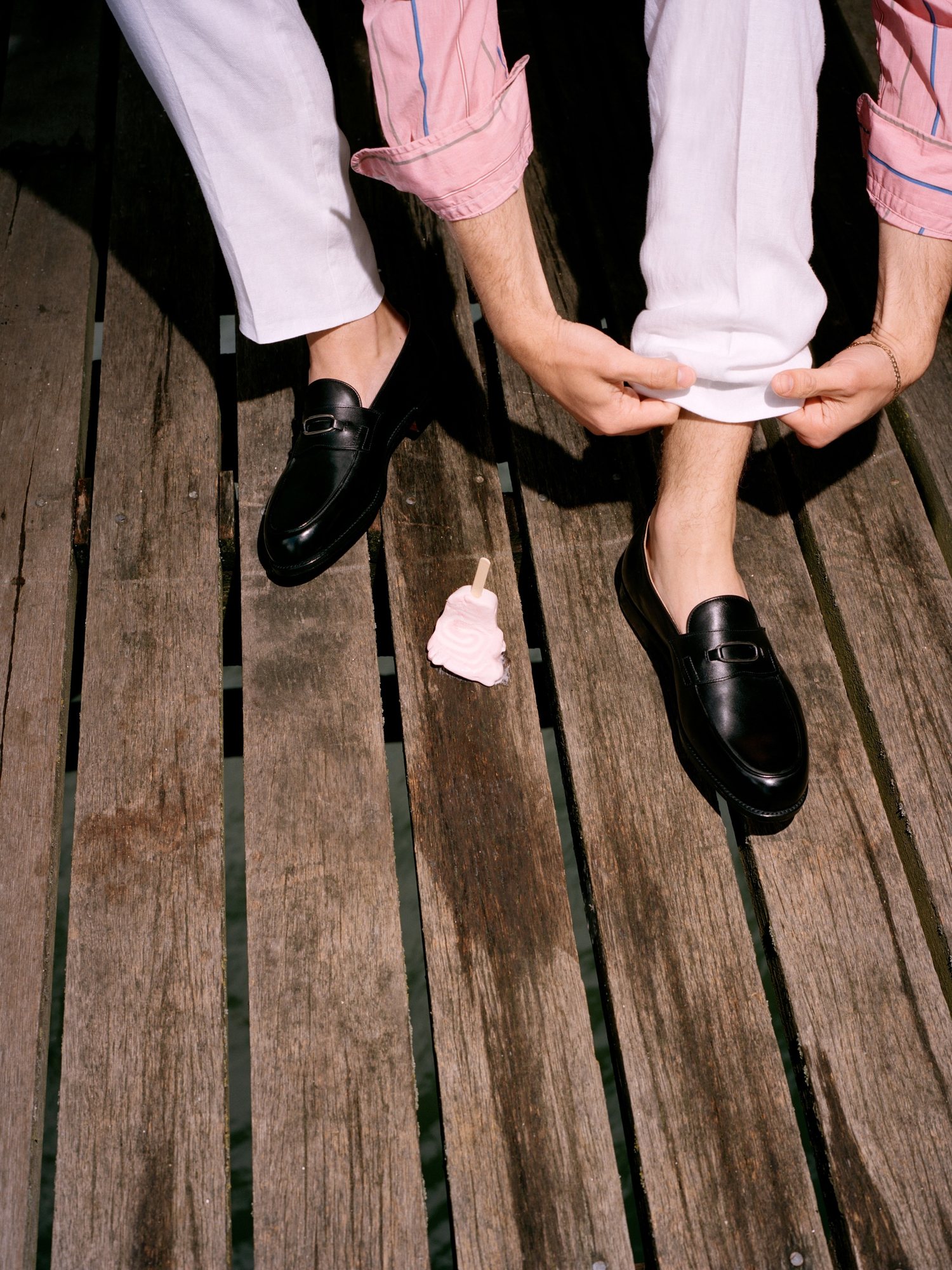 Man in white trousers rolling his cuffs with black loafers on a boardwalk