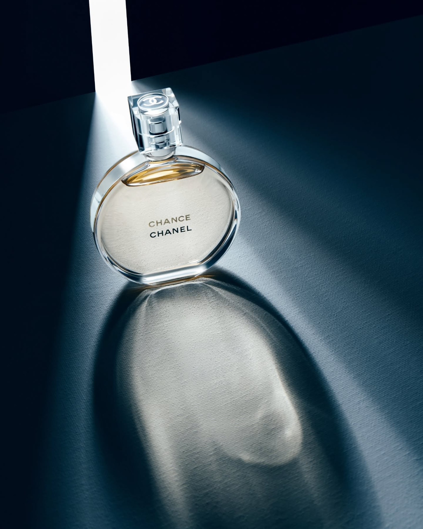 Round bottle of Chanel Chance Eau de Toilette with golden liquid, lit by a beam of light on a dark blue textured surface