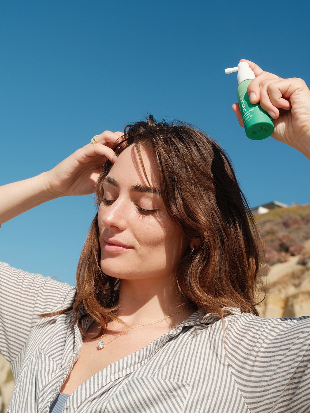 Woman with long hair misting her scalp outdoors beneath a bright blue sky