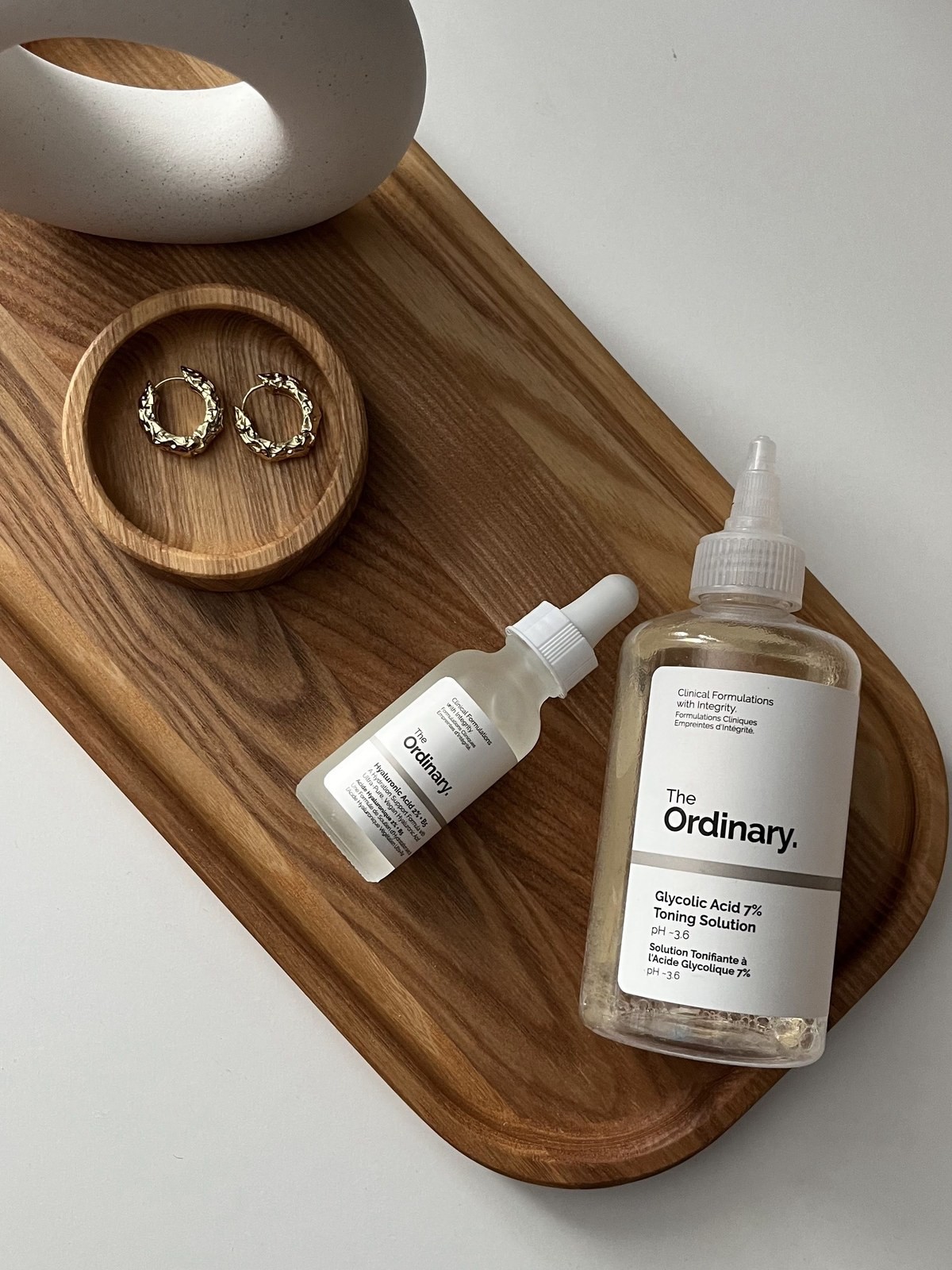 The Ordinary Hyaluronic Acid and Glycolic Acid Toning Solution bottles on a wooden tray beside gold earrings