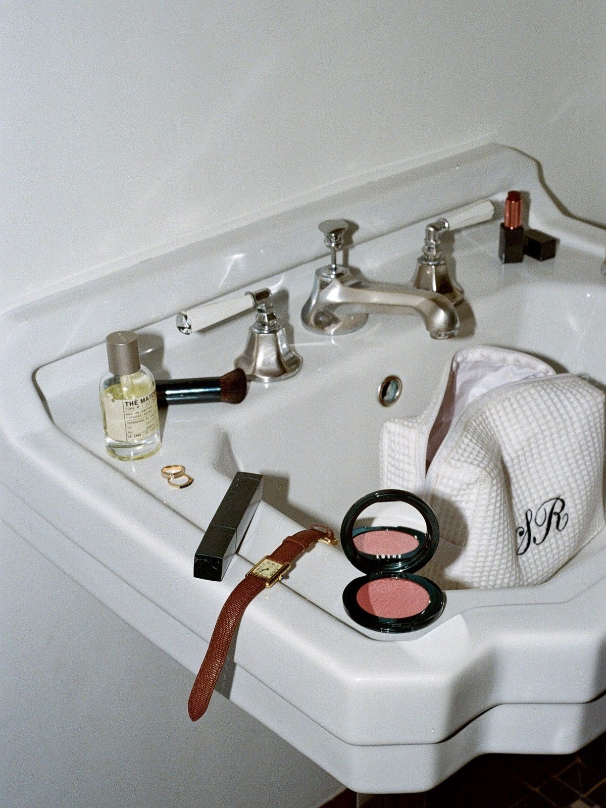 Bathroom sink with a blush compact, lipstick, makeup brush, perfume bottle, gold watch and a monogrammed waffle pouch