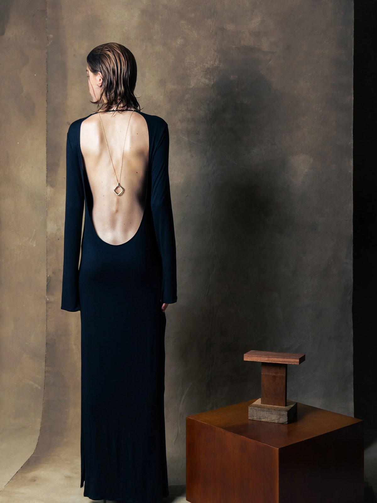 Model in an open-back black gown beside a wooden sculpture in a muted studio