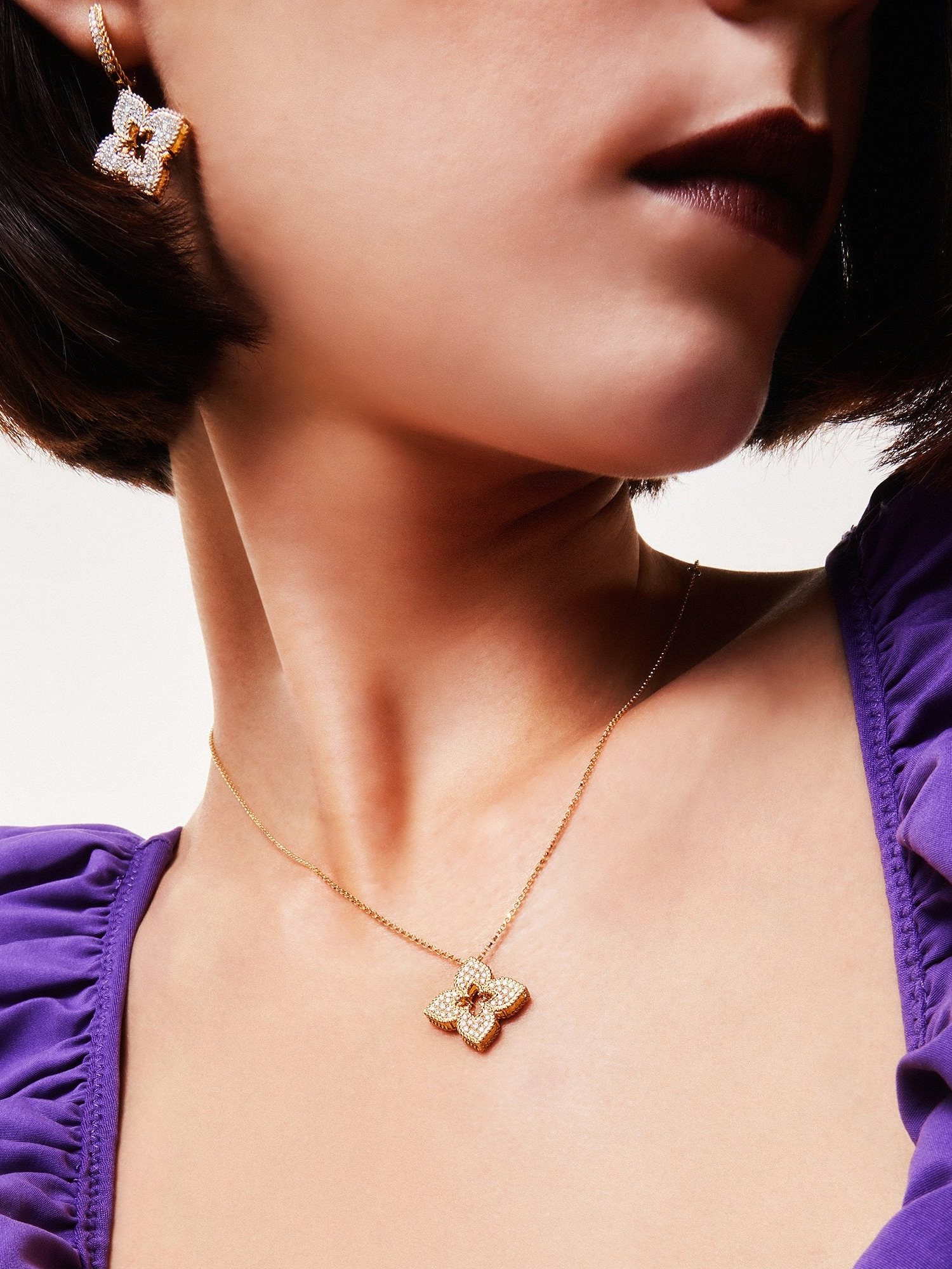 Close up of a model in a purple ruffle top wearing a gold flower pendant and earring