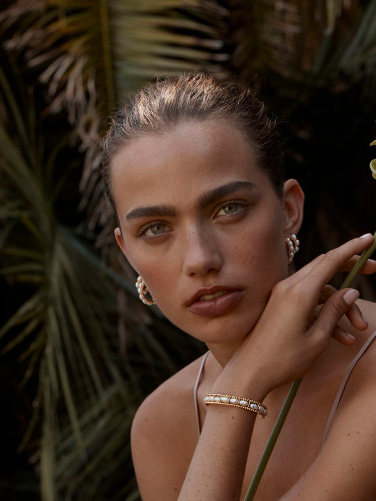 Model holding a tropical flower and wearing pearl and gold jewelry among dark foliage