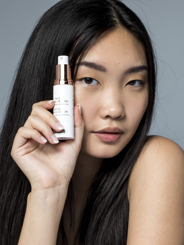 Asian model holds a white Triple Active Retinol bottle beside her face in a gray studio portrait.