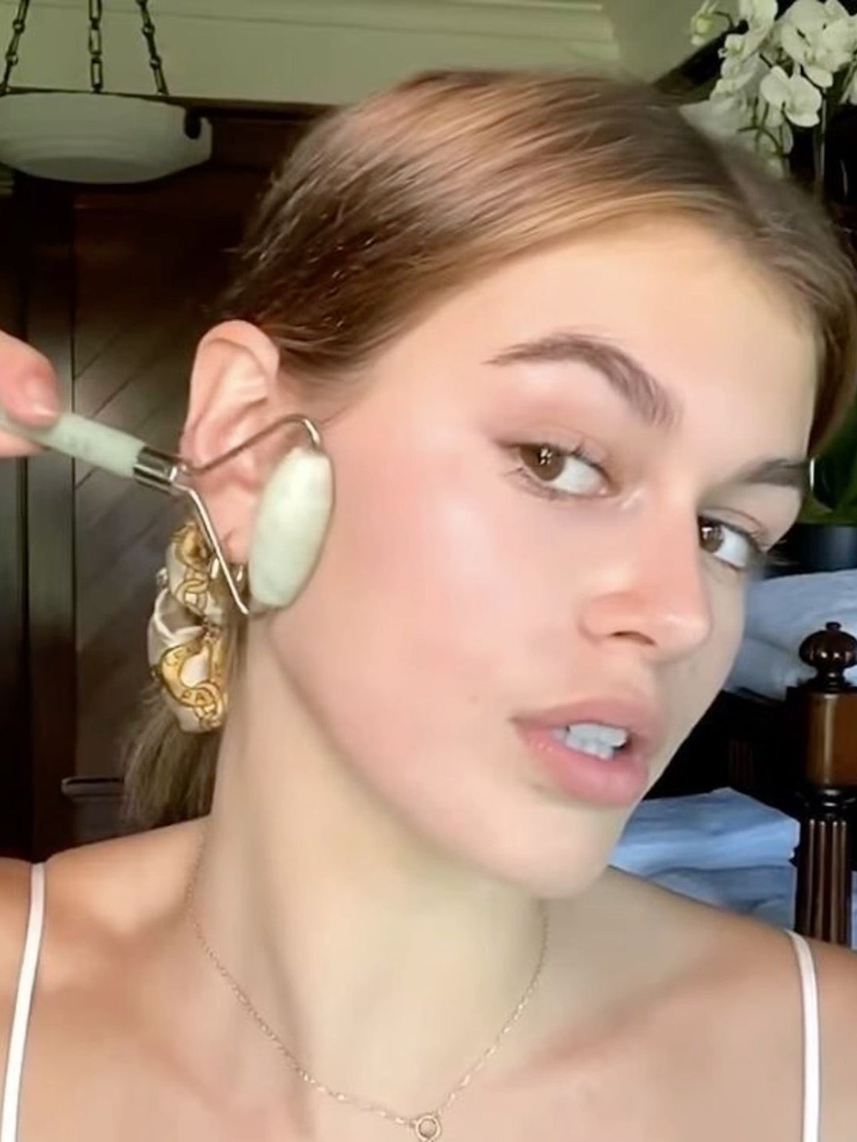 Woman with slicked-back hair rolling a pale jade roller along her jaw near her ear, a silk scarf in her ponytail