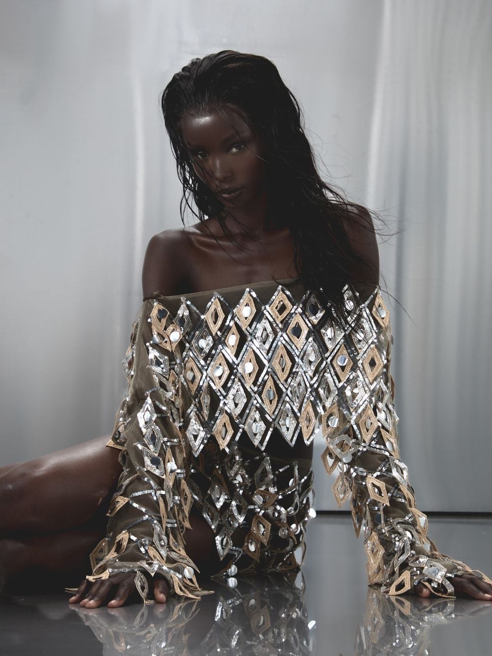 Model in a geometric embellished outfit seated against a pale gray studio backdrop