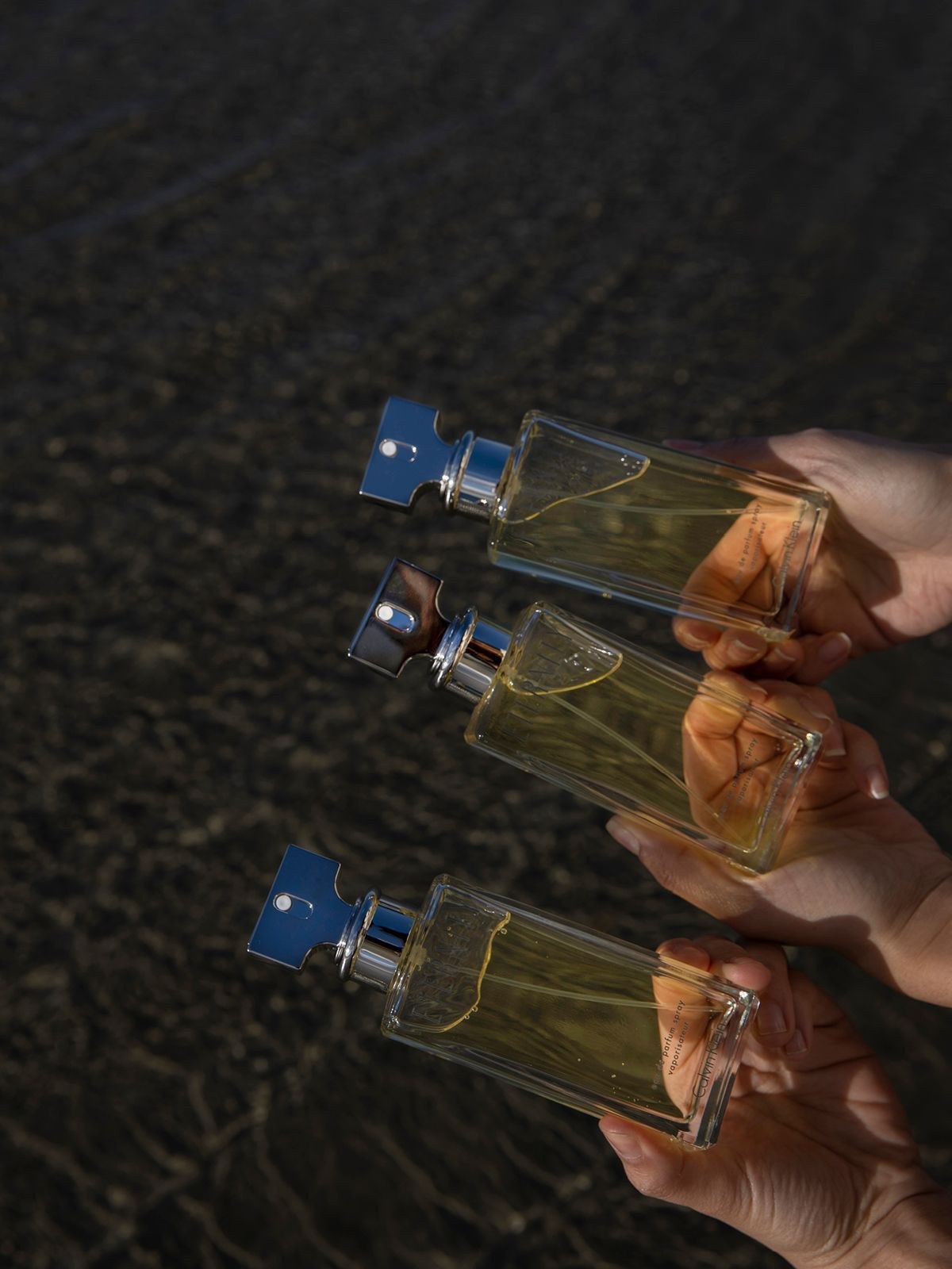 Hands holding three Calvin Klein Eternity bottles with blue and silver caps over dark wet sand