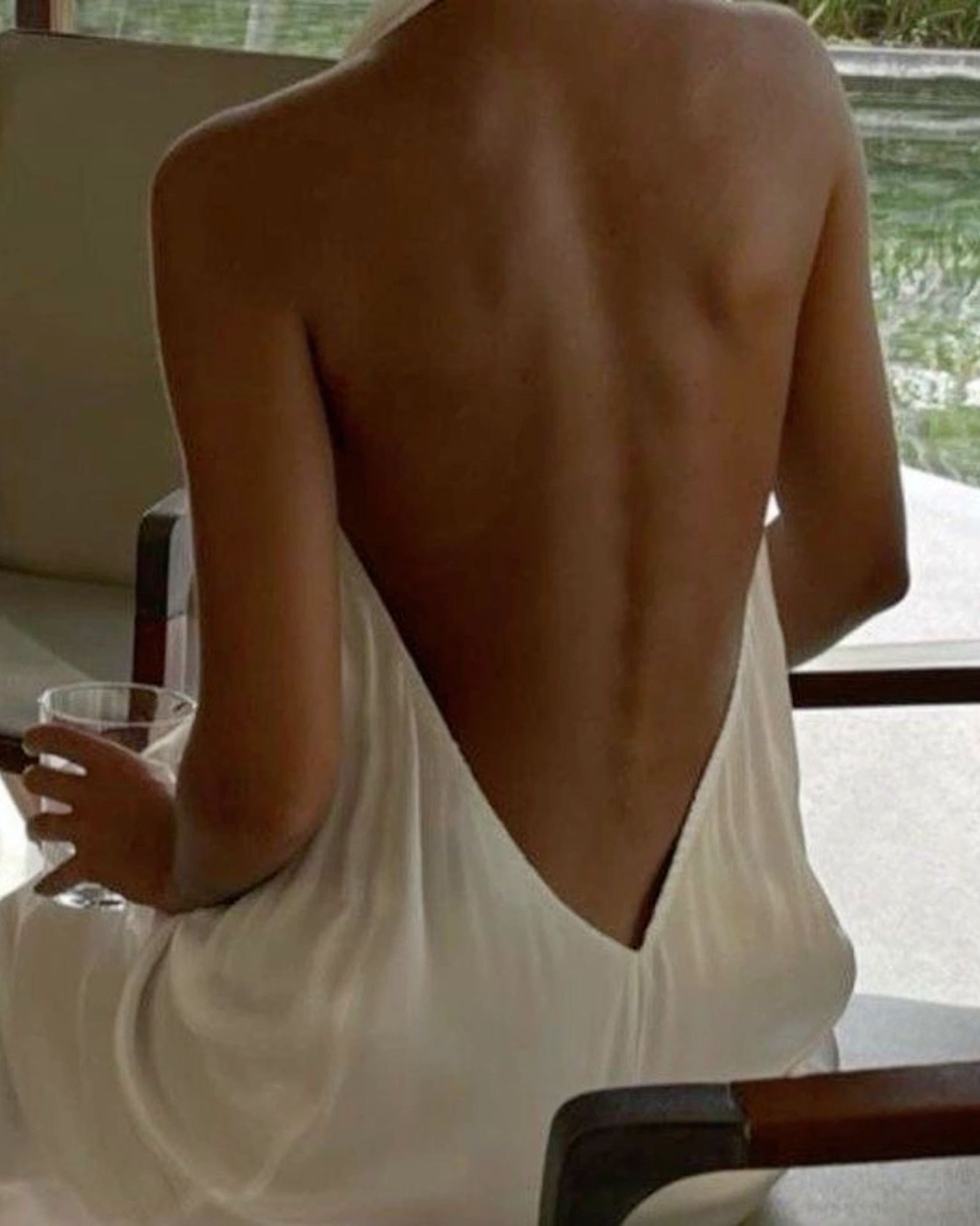 Woman seen from behind in a white low-back slip dress, sitting in a chair holding a glass beside a pool
