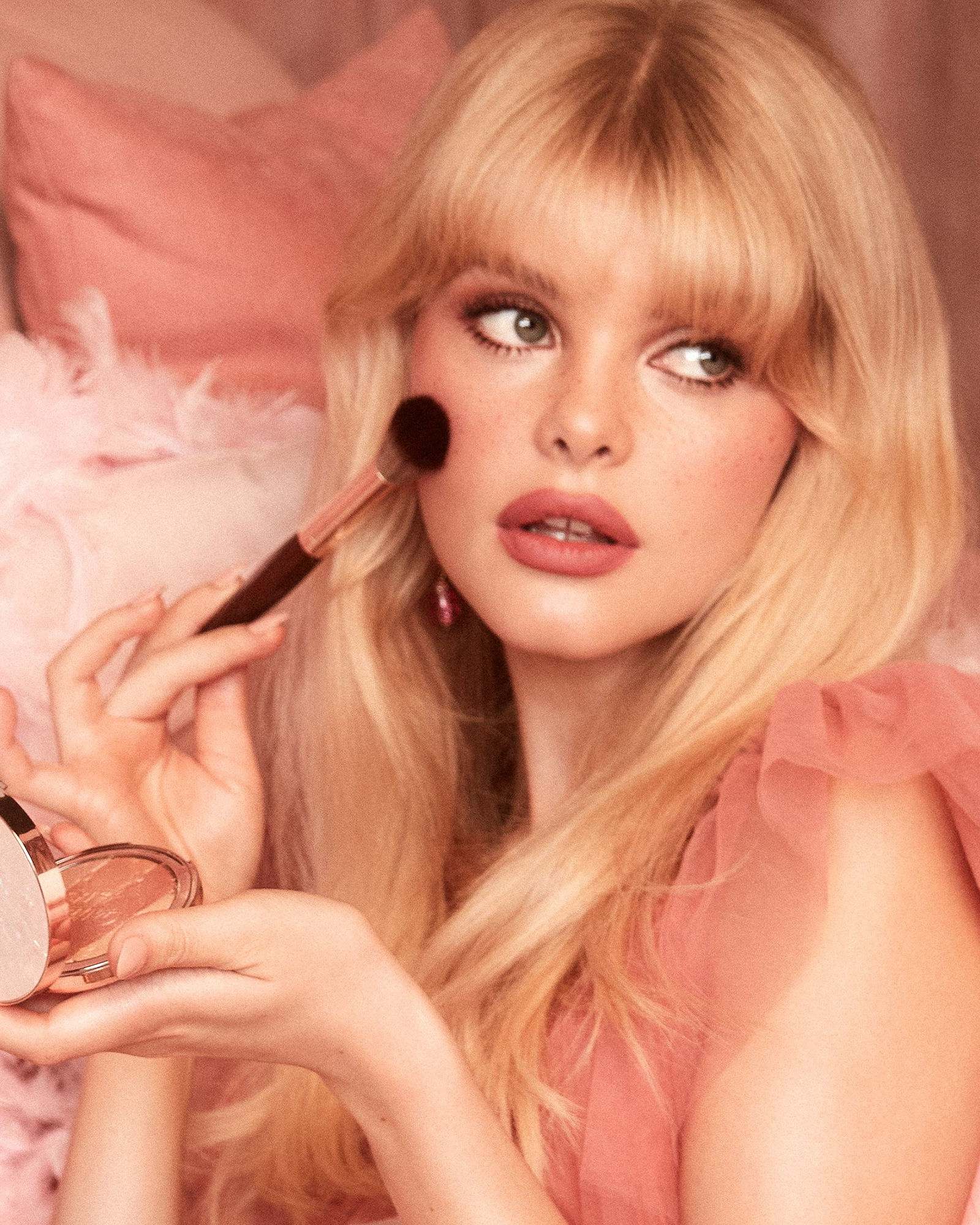 Woman with a blonde fringe and rosy-brown lips brushing blush onto her cheek, wearing a coral tulle dress against pink pillows