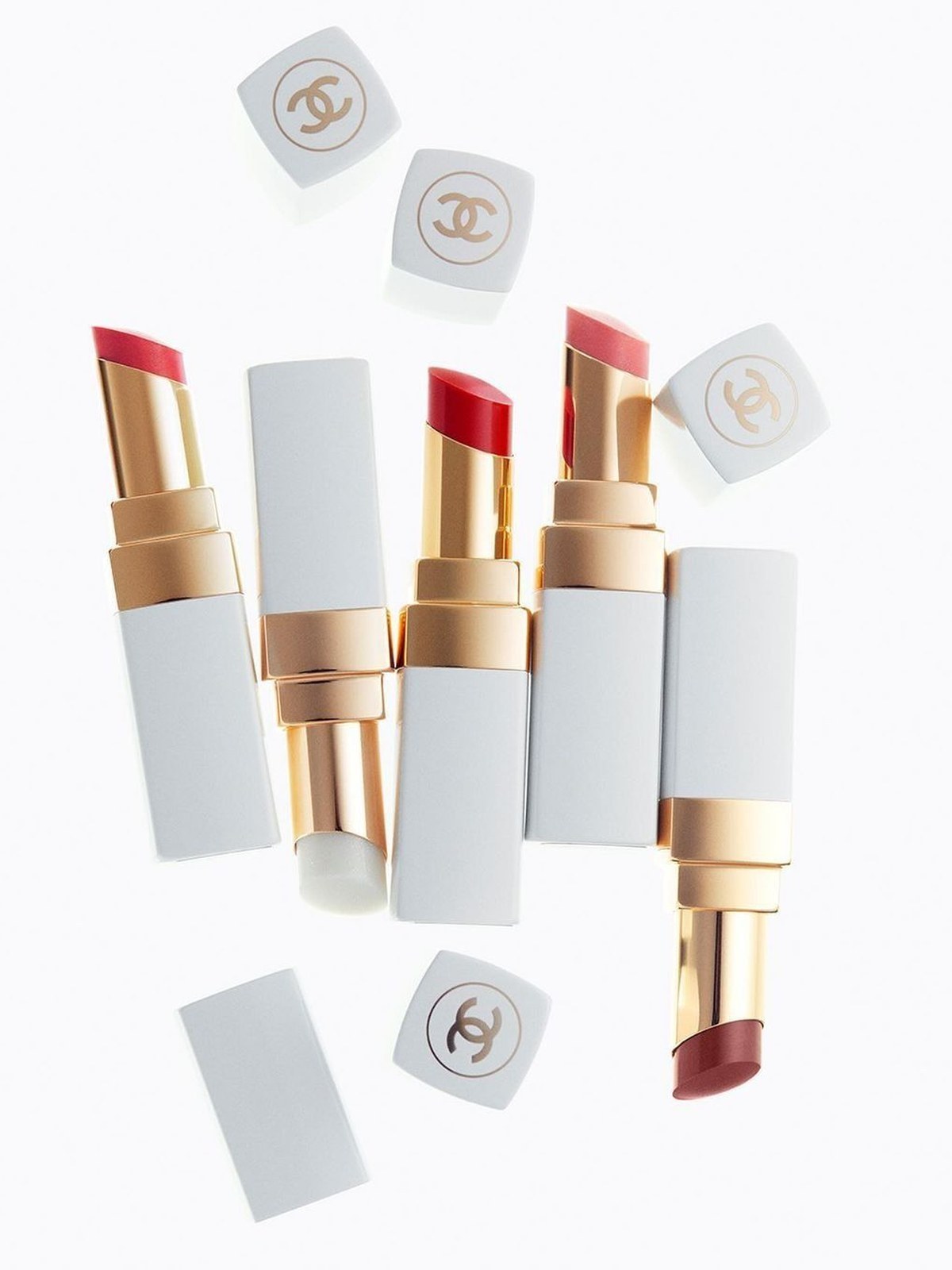Five open Chanel lip balms in white and gold tubes in red, coral and rose, scattered on a white background