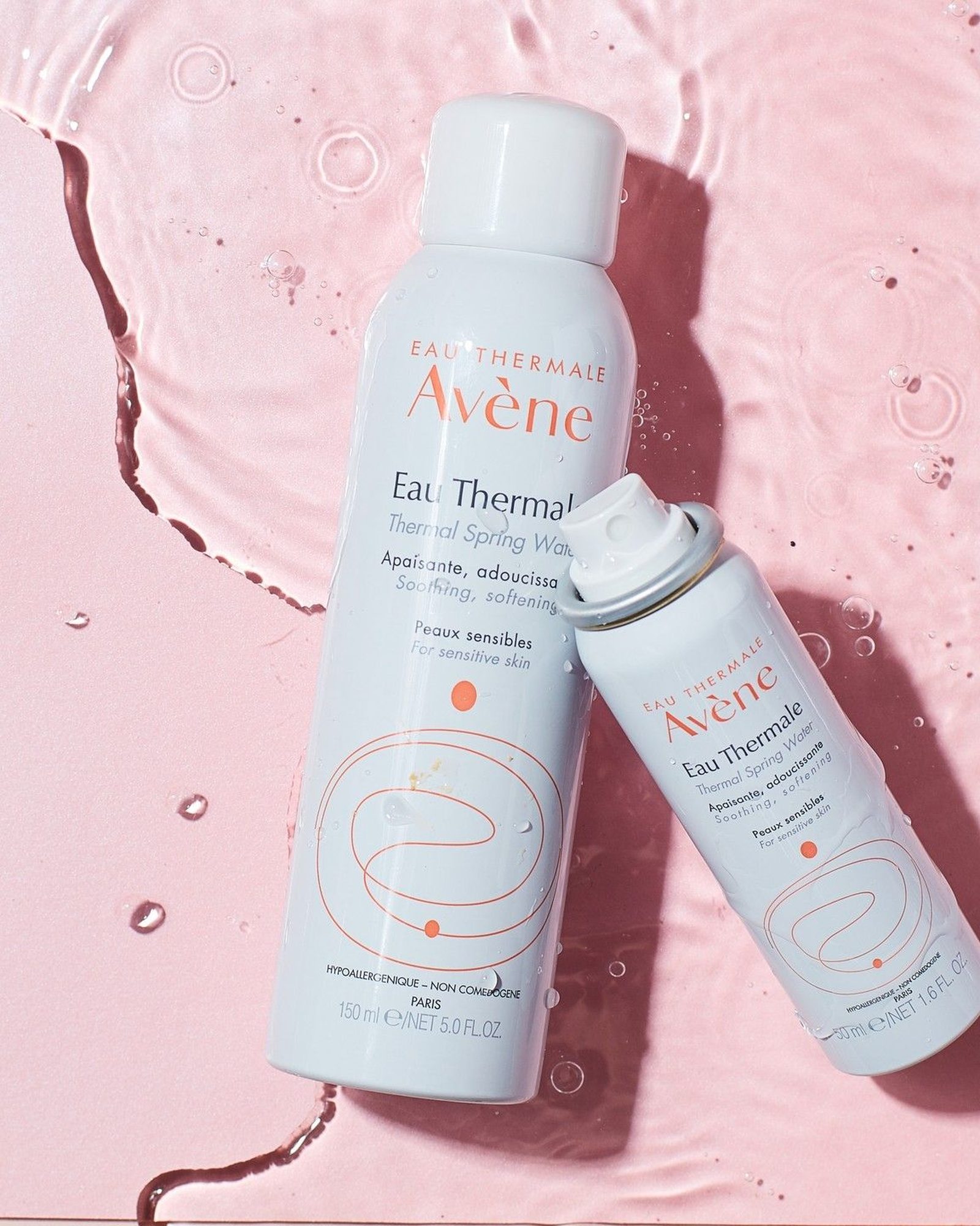 Two Eau Thermale Avène Thermal Spring Water spray cans, one large and one travel size, lying in pink water with ripples and droplets