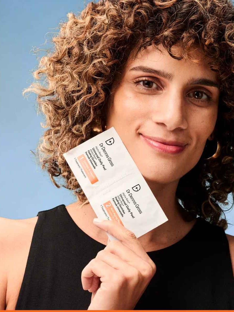 Curly-haired woman holding Alpha Beta peel packets against a blue backdrop