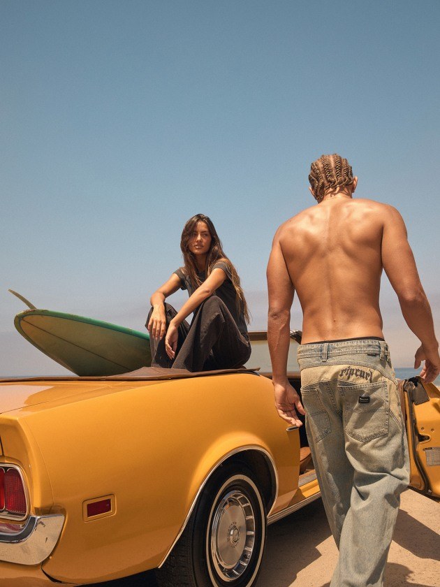 A woman in the Abrand and Rip Curl collaboration sits near a beach with a second model and a vintage car