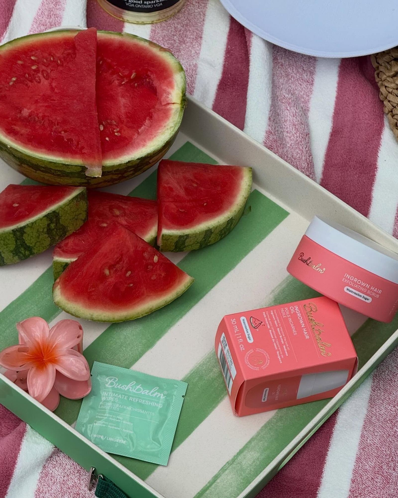 Sliced watermelon on a green striped tray beside coral Bushbalm oil and scrub, a wipe packet and a pink flower claw clip on a pink striped towel