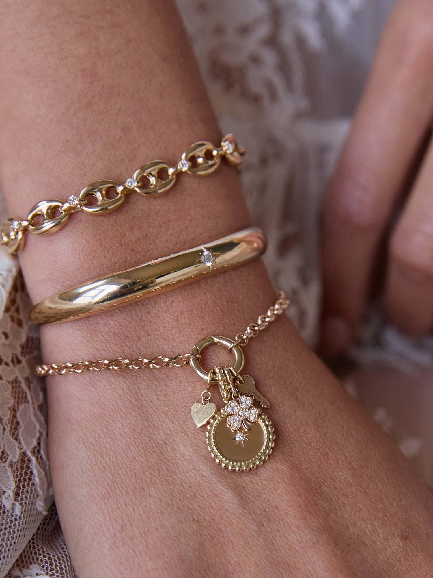 Wrist with stacked gold bracelets and a charm bracelet against lace