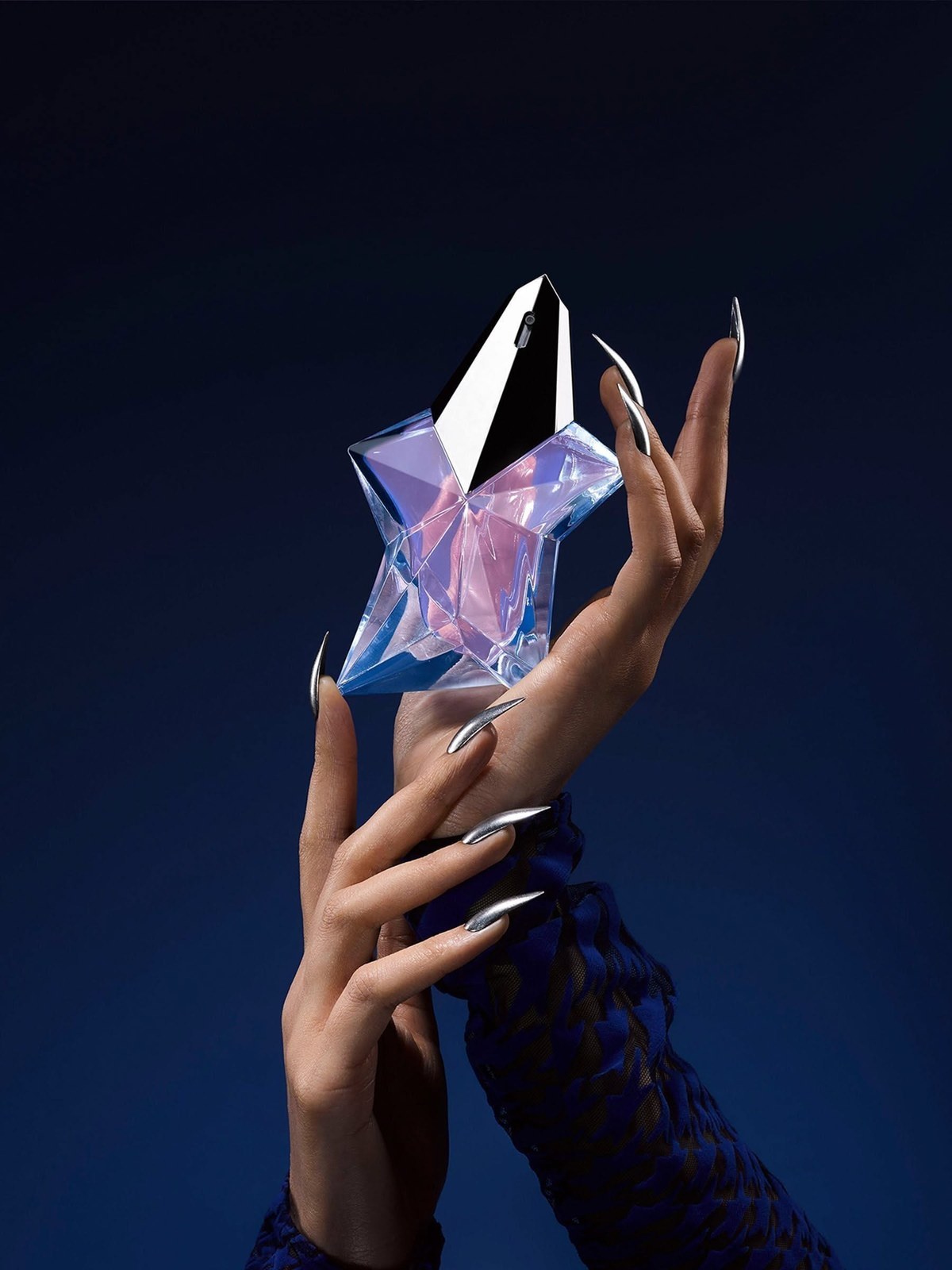 Hands with long silver nails holding a star shaped Mugler perfume bottle against a deep blue background