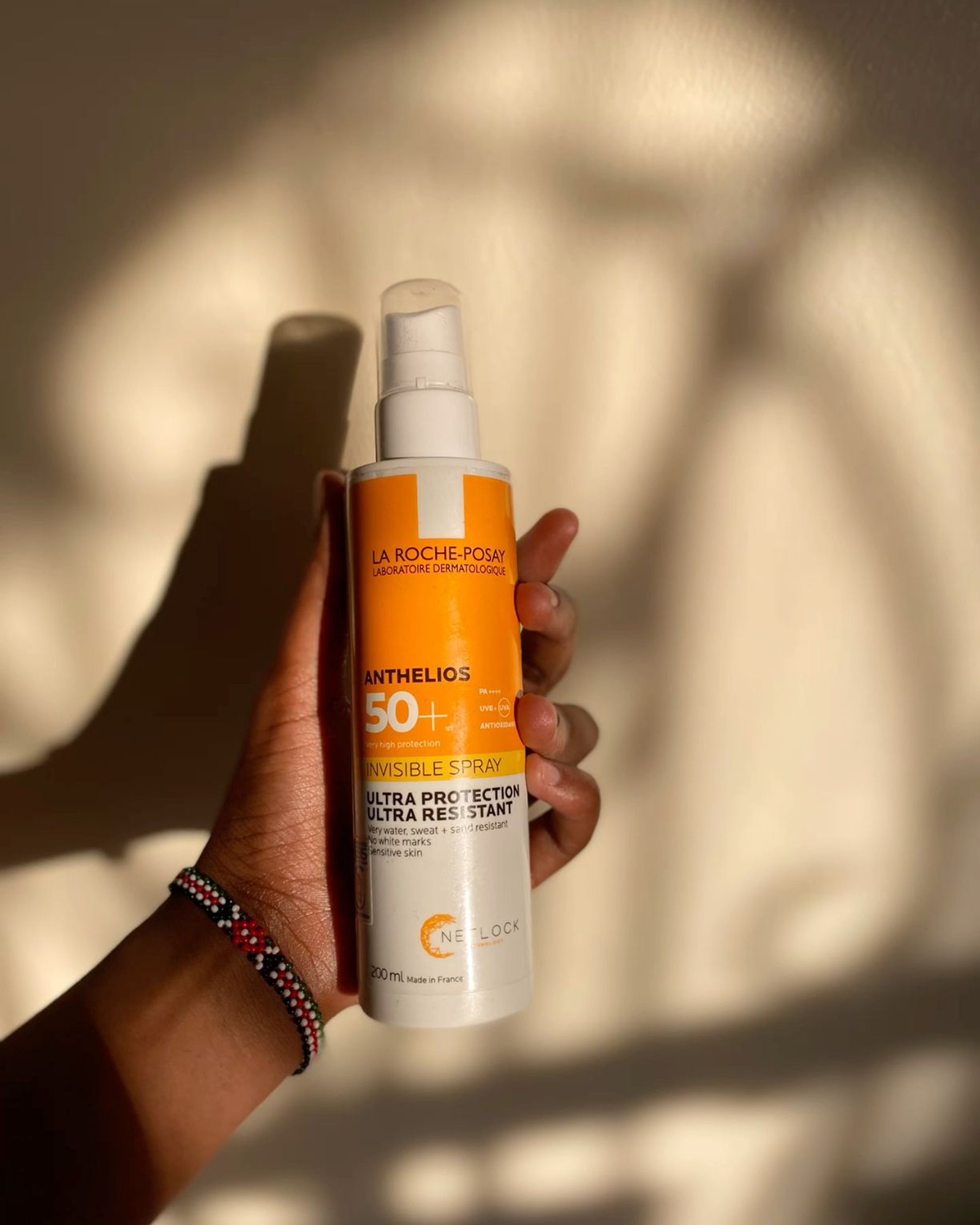 Hand holding a La Roche-Posay Anthelios Invisible Spray SPF 50+ bottle against a sunlit wall with soft shadows