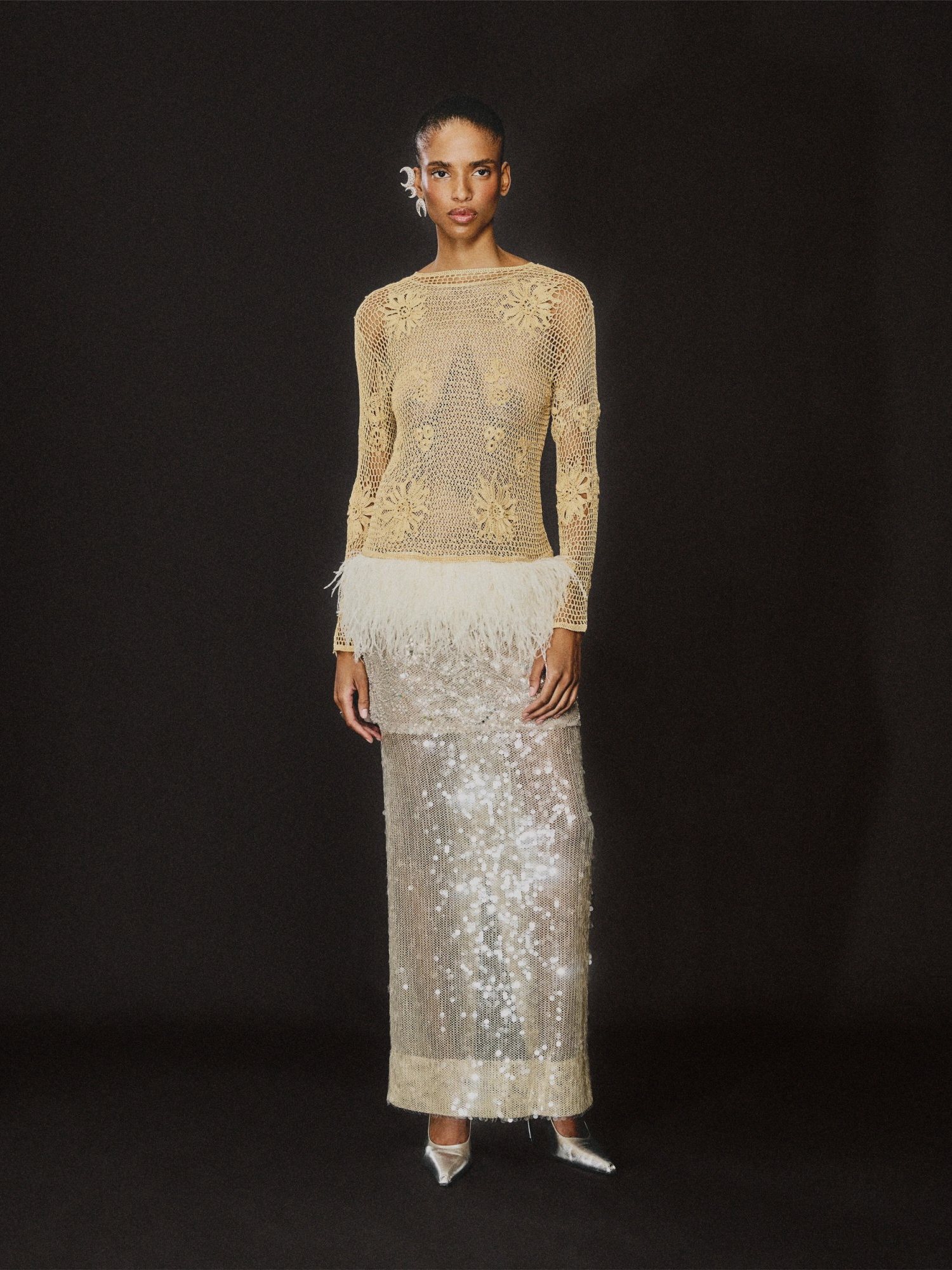 Model in a sheer embroidered top and sequined column skirt on a black backdrop