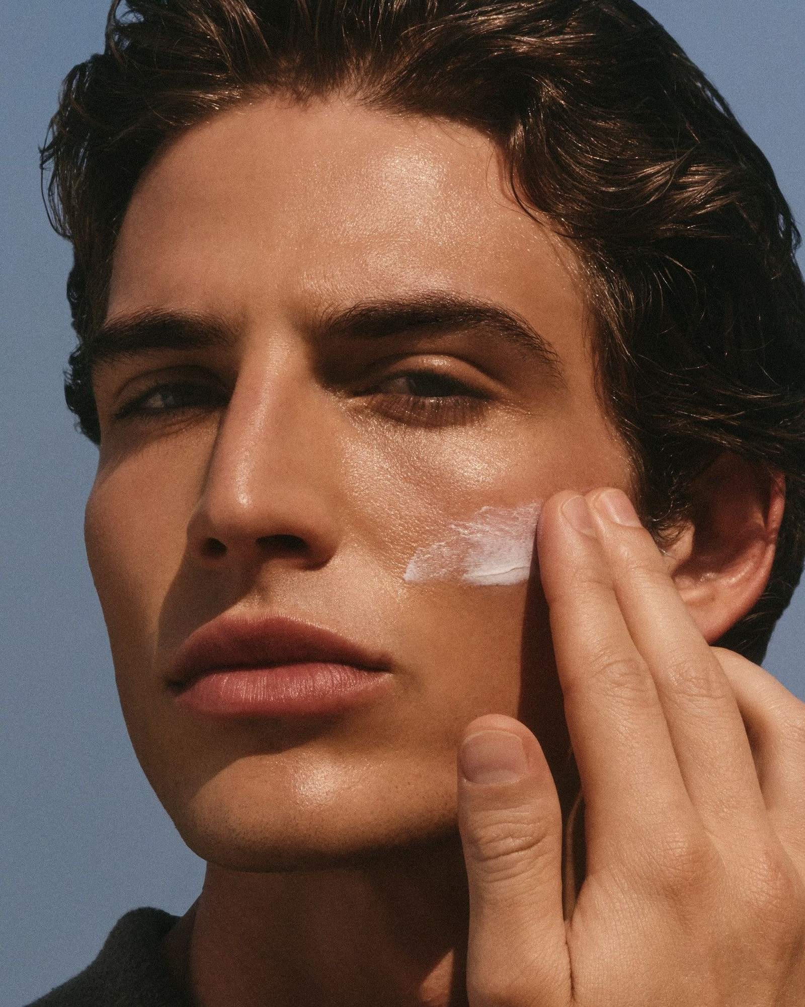 Man with slicked dark hair smoothing a streak of white cream onto his cheek against a blue sky