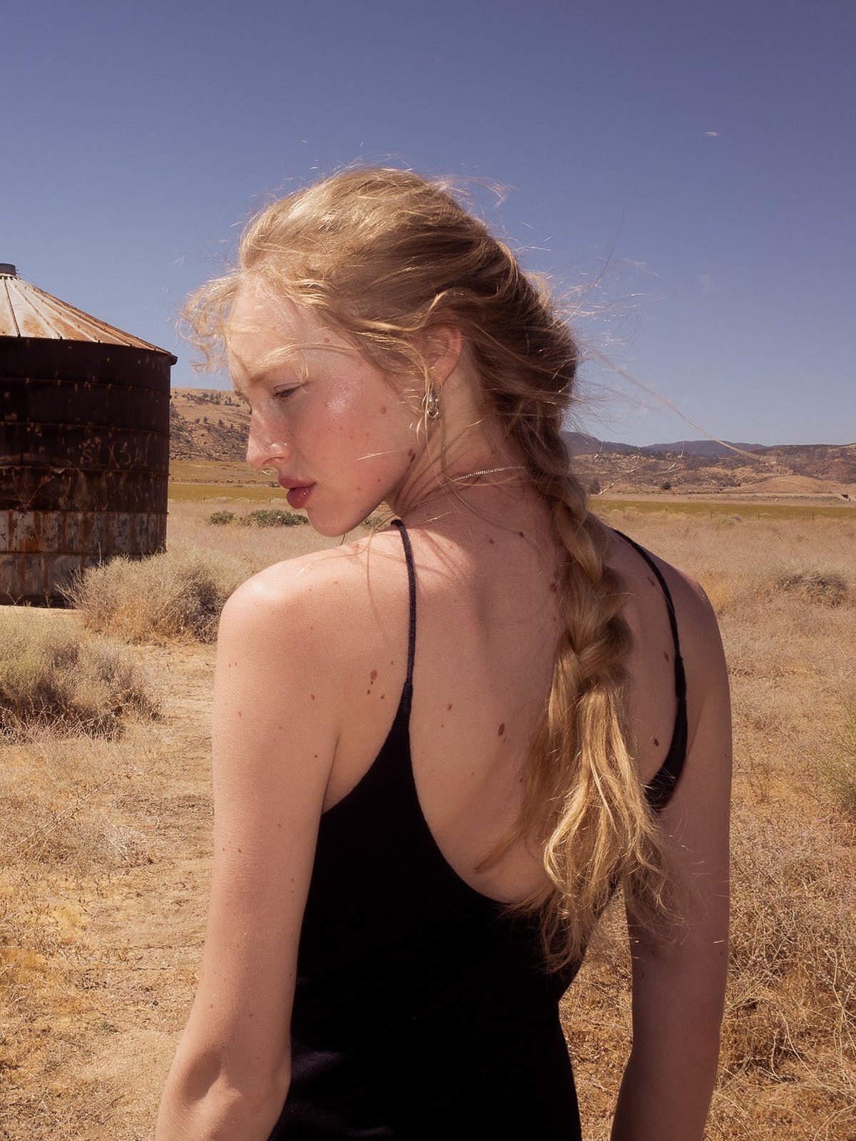 Model in a black camisole with a side braid in front of rusted grain silos