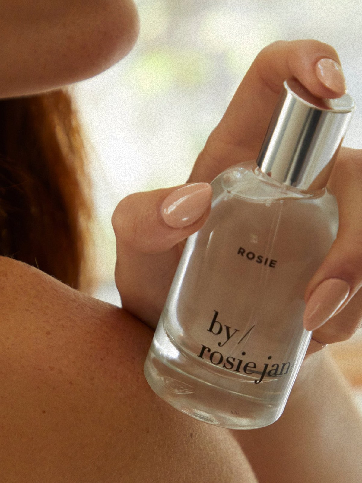 Close-up of a person applying Rosie perfume to their shoulder in natural light