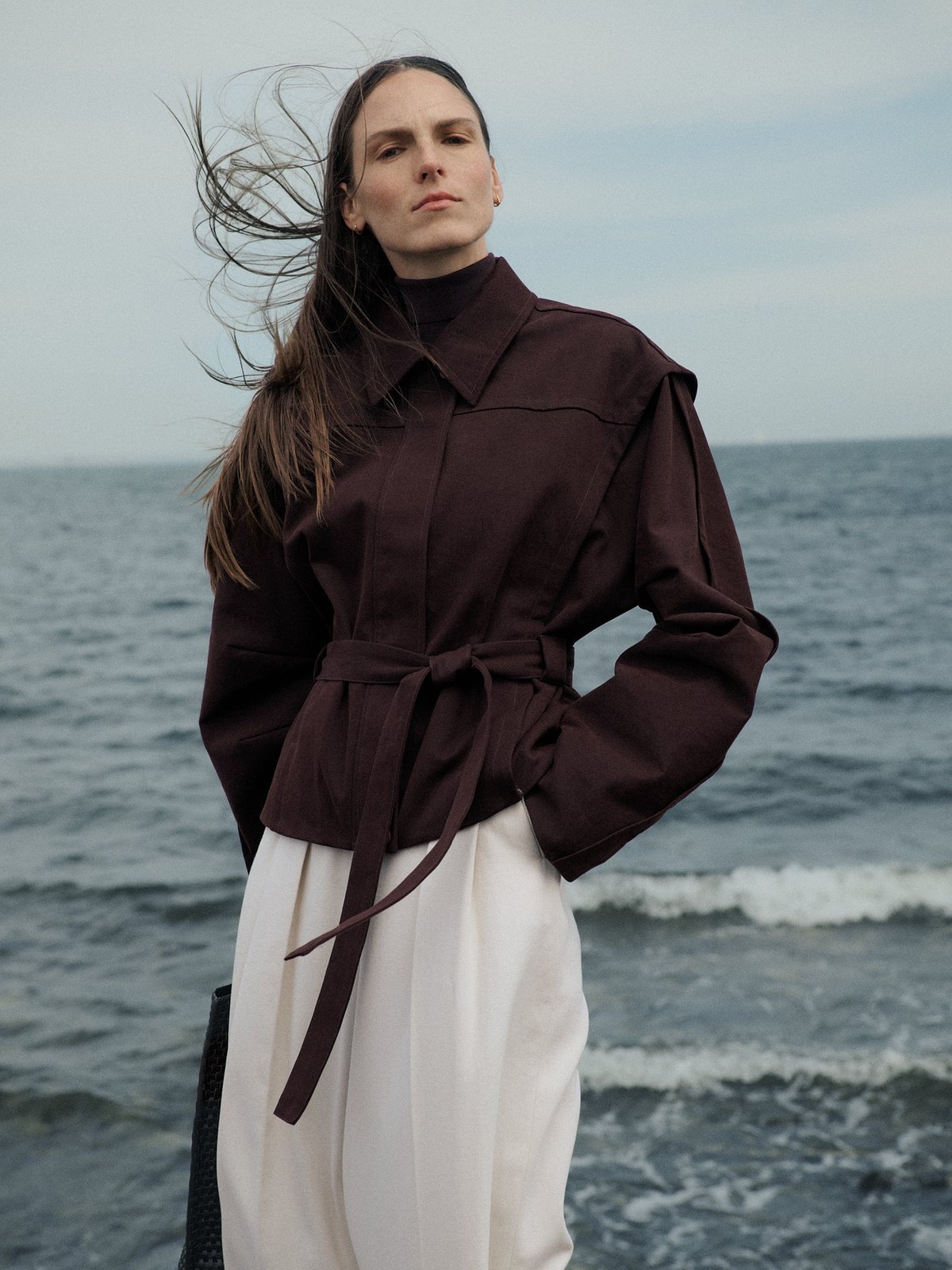 Model in a dark belted jacket and white trousers by the shore