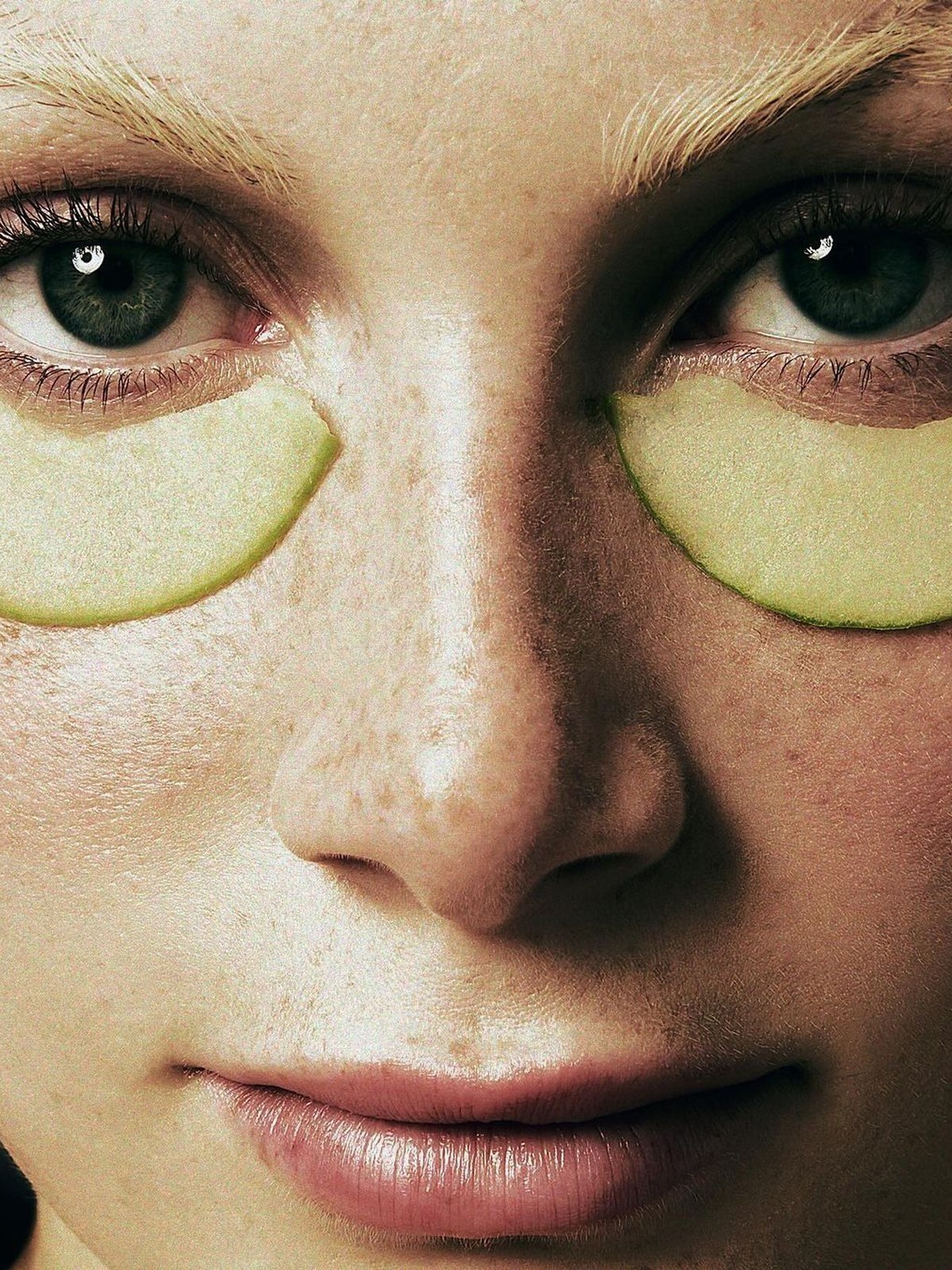 Extreme close-up of a freckled face with pale green eyes and pale green gel patches under each eye