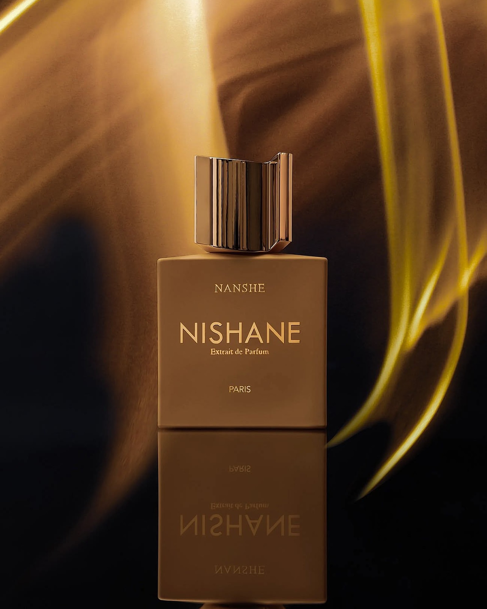 Gold Nishane Nanshe Extrait de Parfum bottle with a reflection against a dark amber background with streaks of golden light