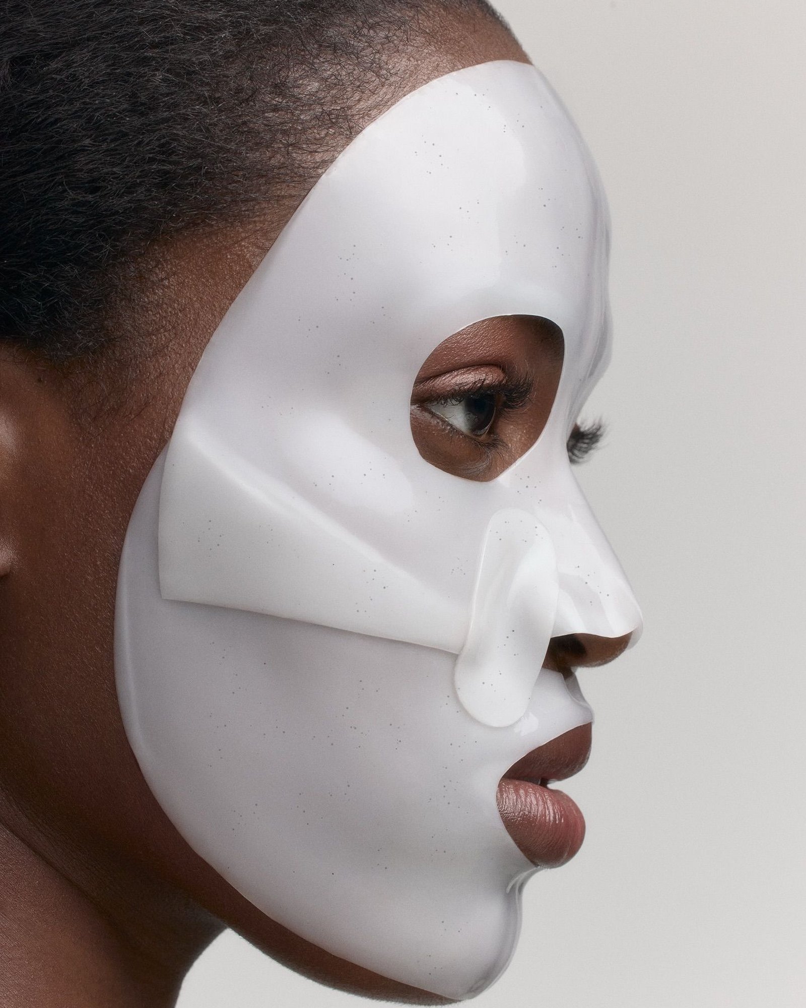 Side profile of a woman wearing a smooth white gel face mask with cut-outs for the eye and nostril against a pale grey background