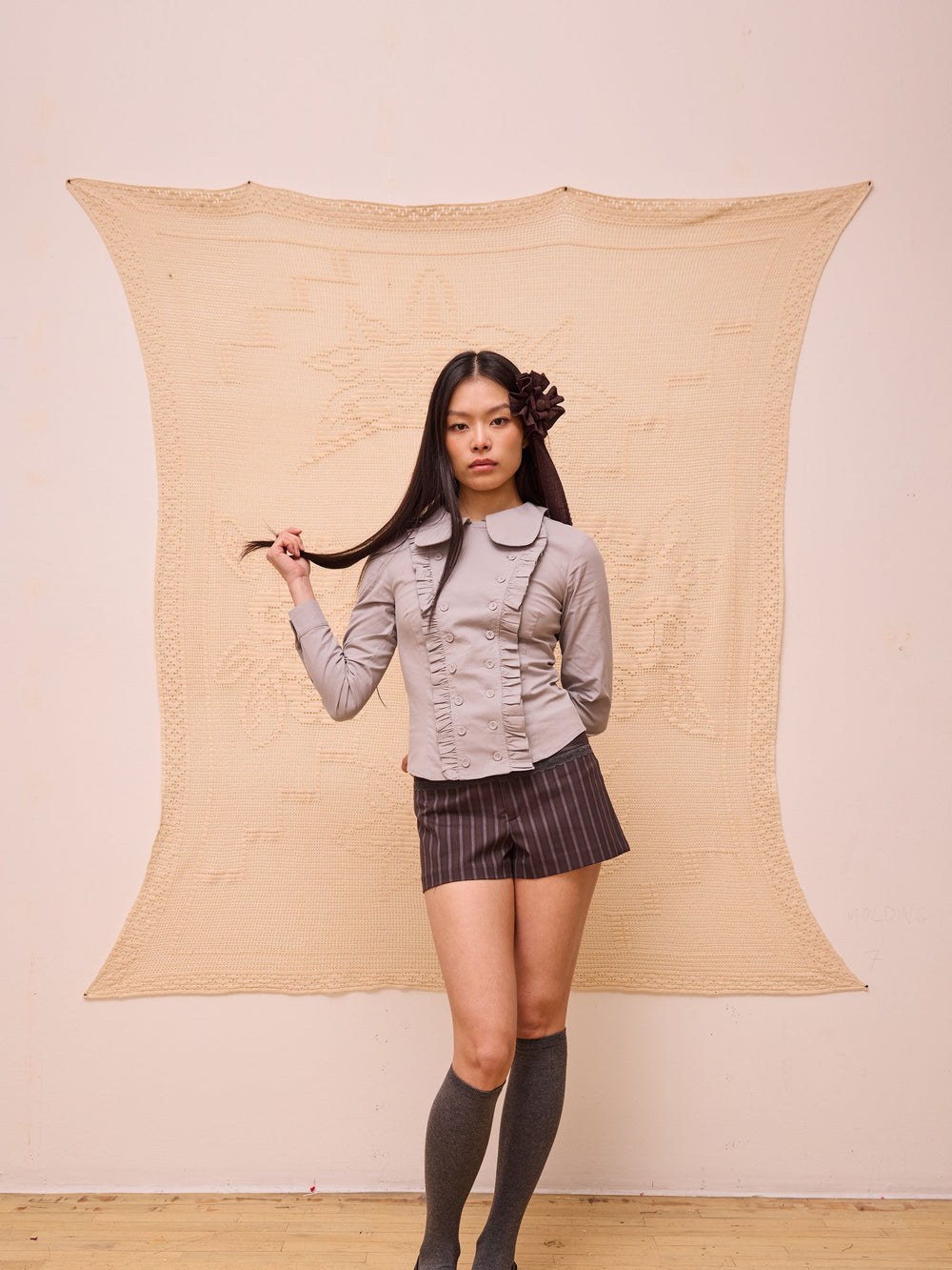 Model in a pale ruffled blouse and pleated shorts against a textured studio backdrop
