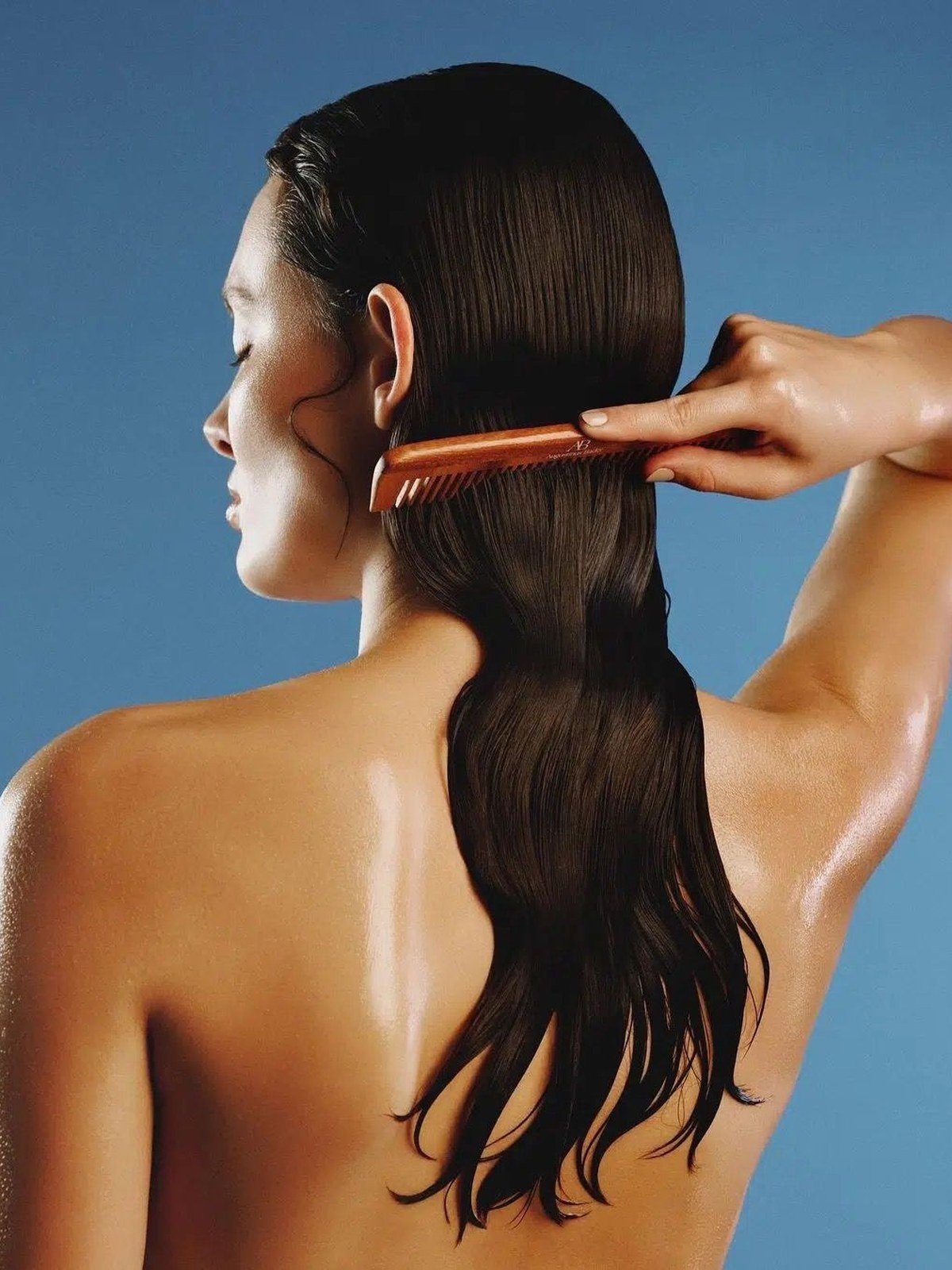 Back view of a woman against a blue background combing long wet dark brown hair with a wooden comb