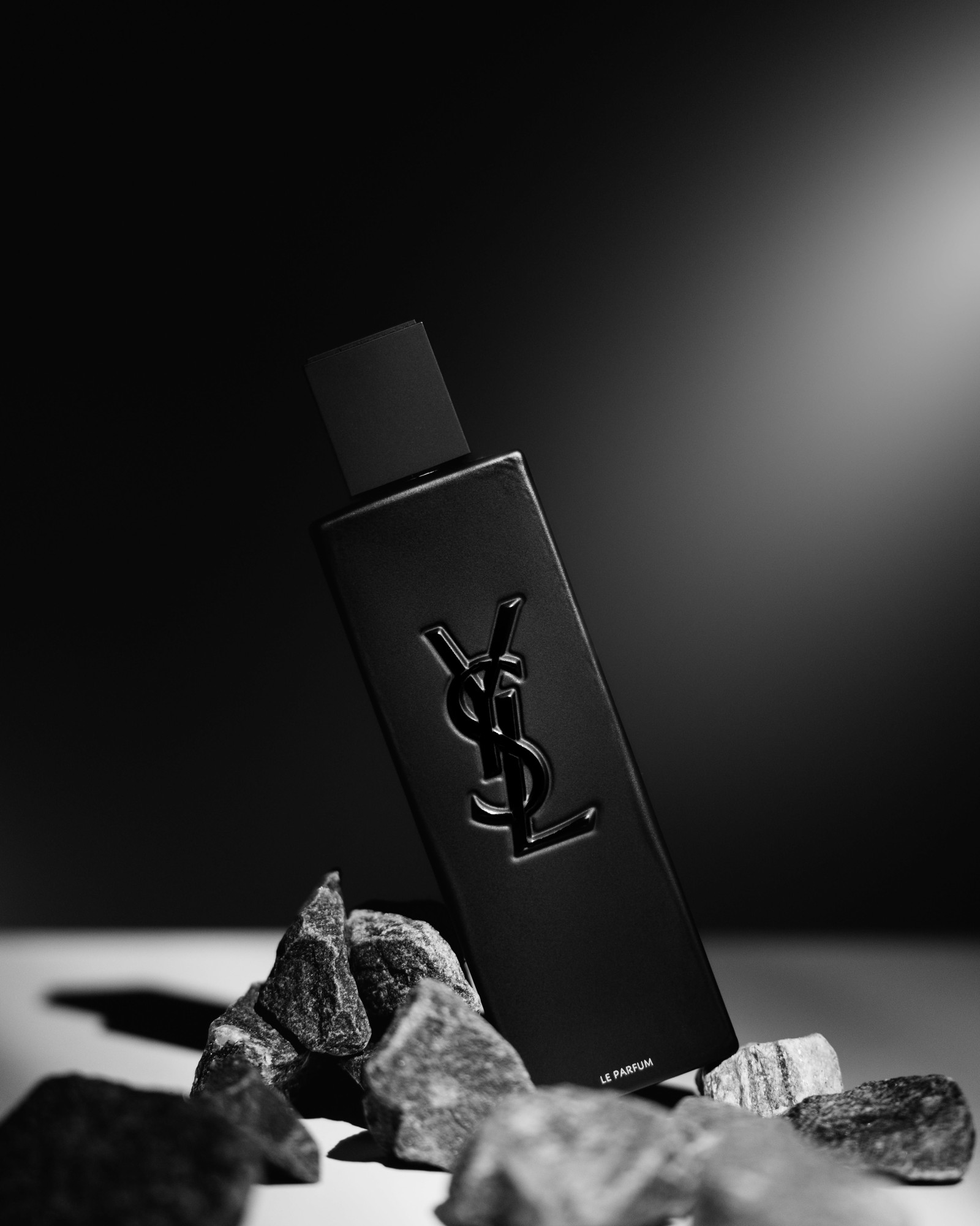 Black and white photo of a black YSL MYSLF Le Parfum bottle leaning on grey rocks in a beam of light