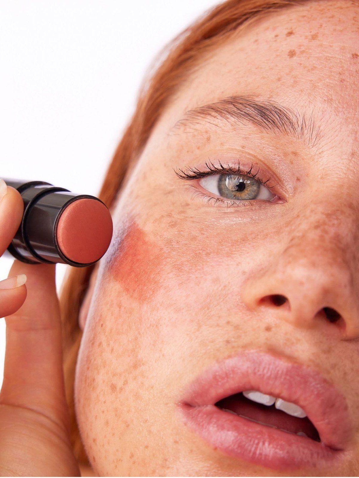 Close-up of a freckled woman holding a terracotta cream blush stick beside a swipe of blush on her cheek