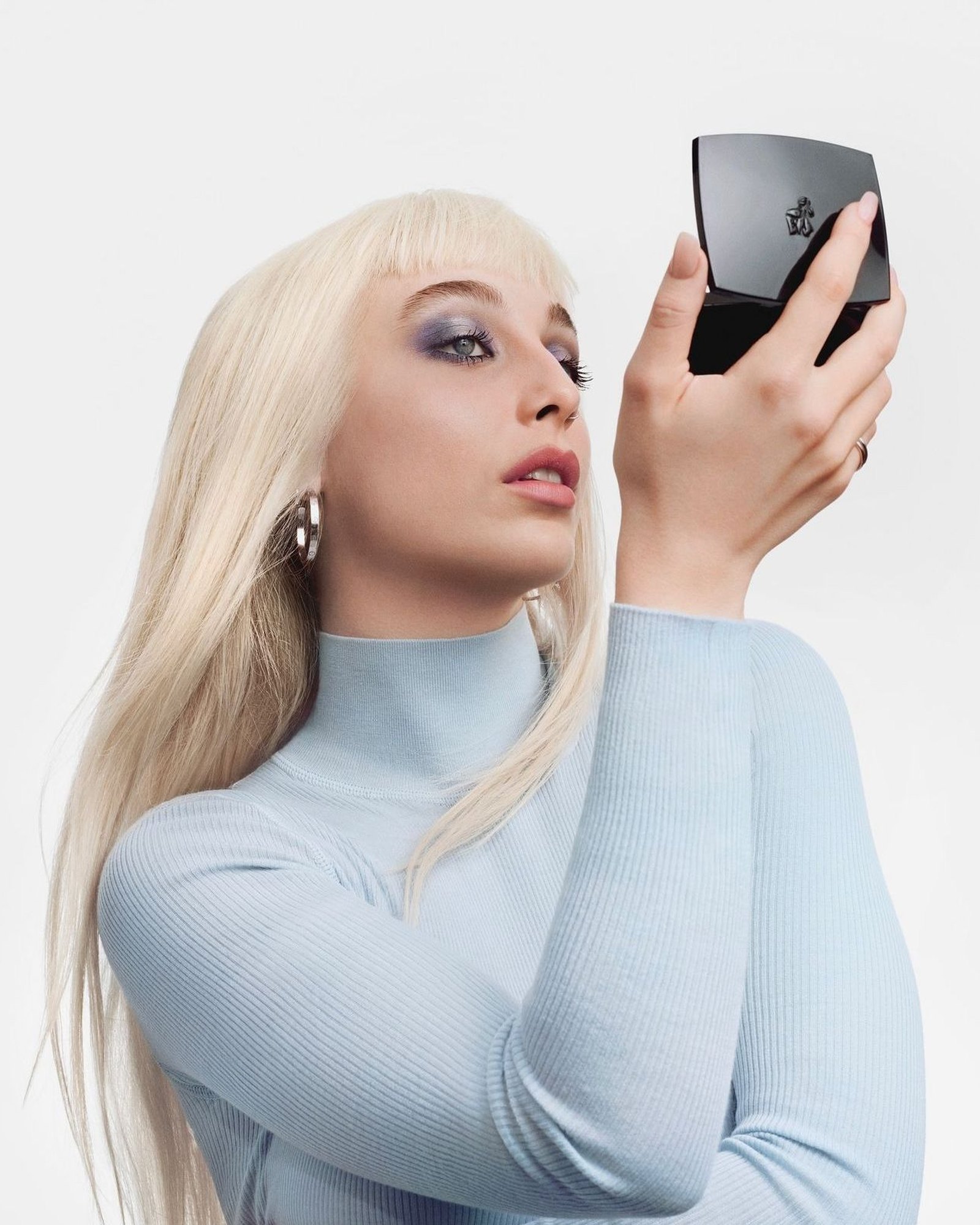 Woman with long platinum blonde hair and bangs in a pale blue ribbed turtleneck holding up a black Lancôme compact like a mirror