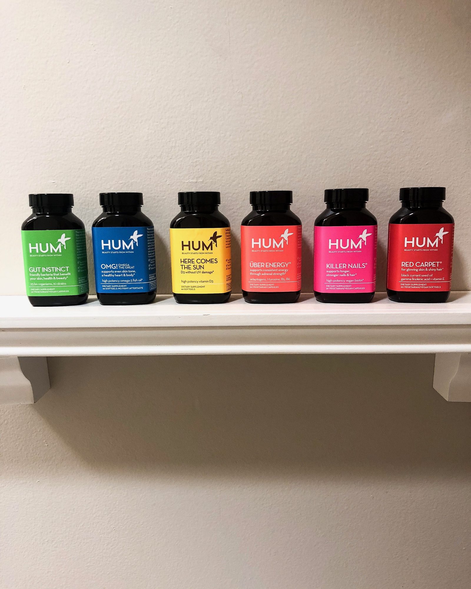 Row of six colorful HUM bottles in green, blue, yellow, orange, pink and red on a white shelf against a cream wall