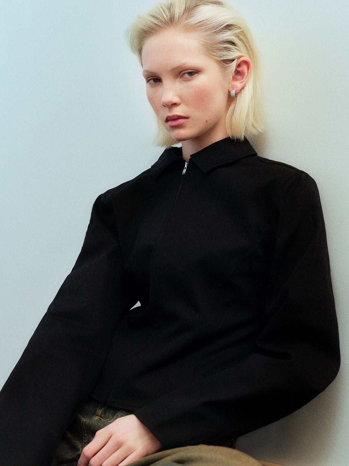 Blonde model in a black zip top seated against a pale blue wall