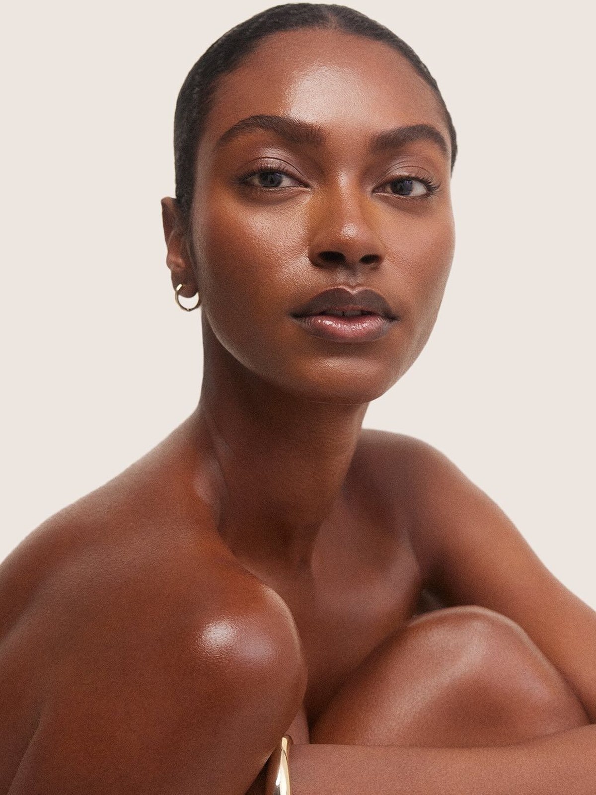 Woman with a deep skin tone and hair pulled back, glossy skin and a gold cuff, sitting with her arms around her knees against a pale backdrop