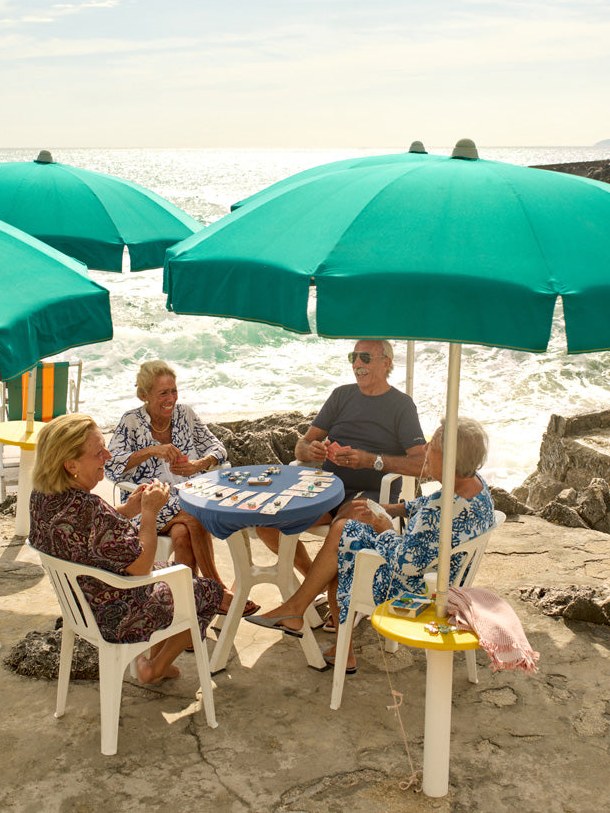 Friends playing cards beneath turquoise umbrellas at a rocky Italian beach club.