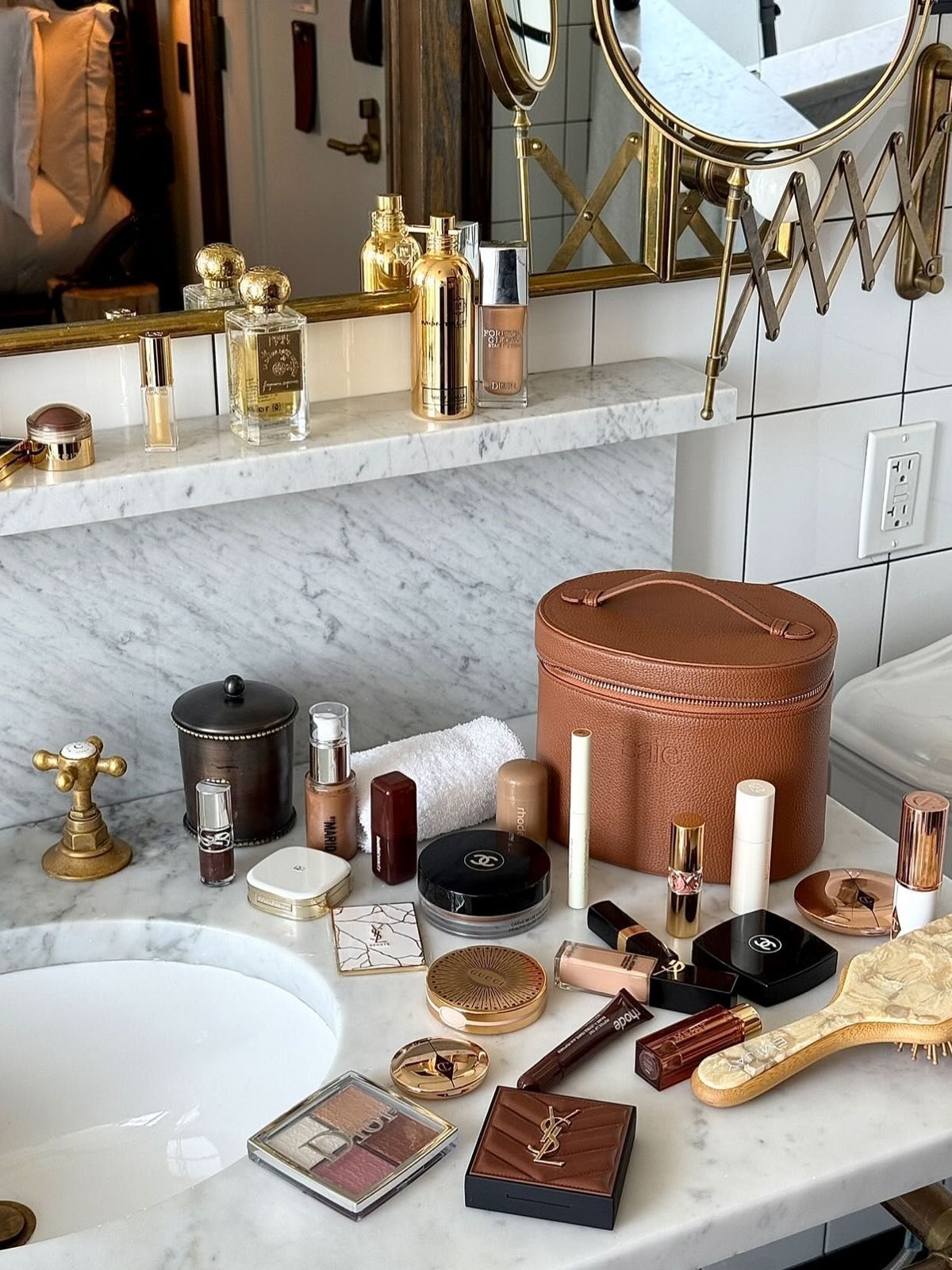 Marble bathroom vanity covered in makeup compacts, lipsticks and perfumes beside a tan leather round beauty case