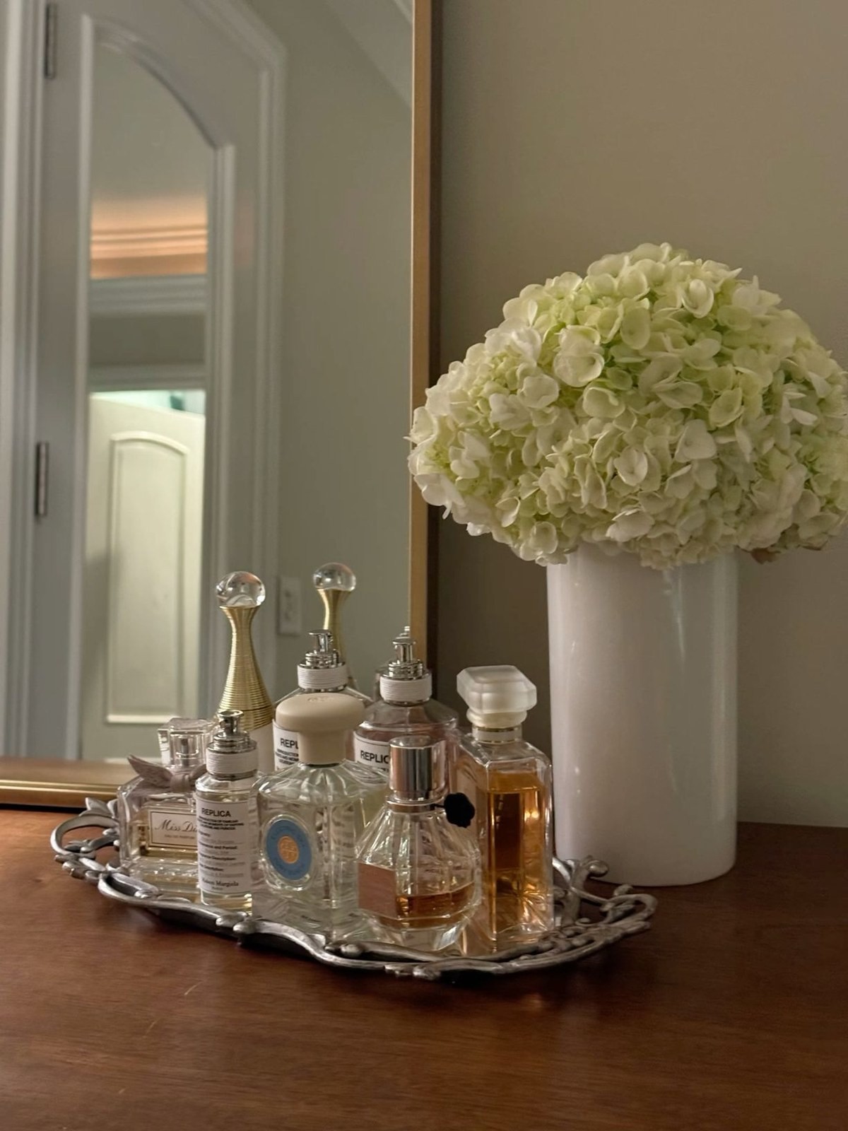 Perfume bottles including Dior and Maison Margiela Replica lined up on a mirrored tray beside white hydrangeas
