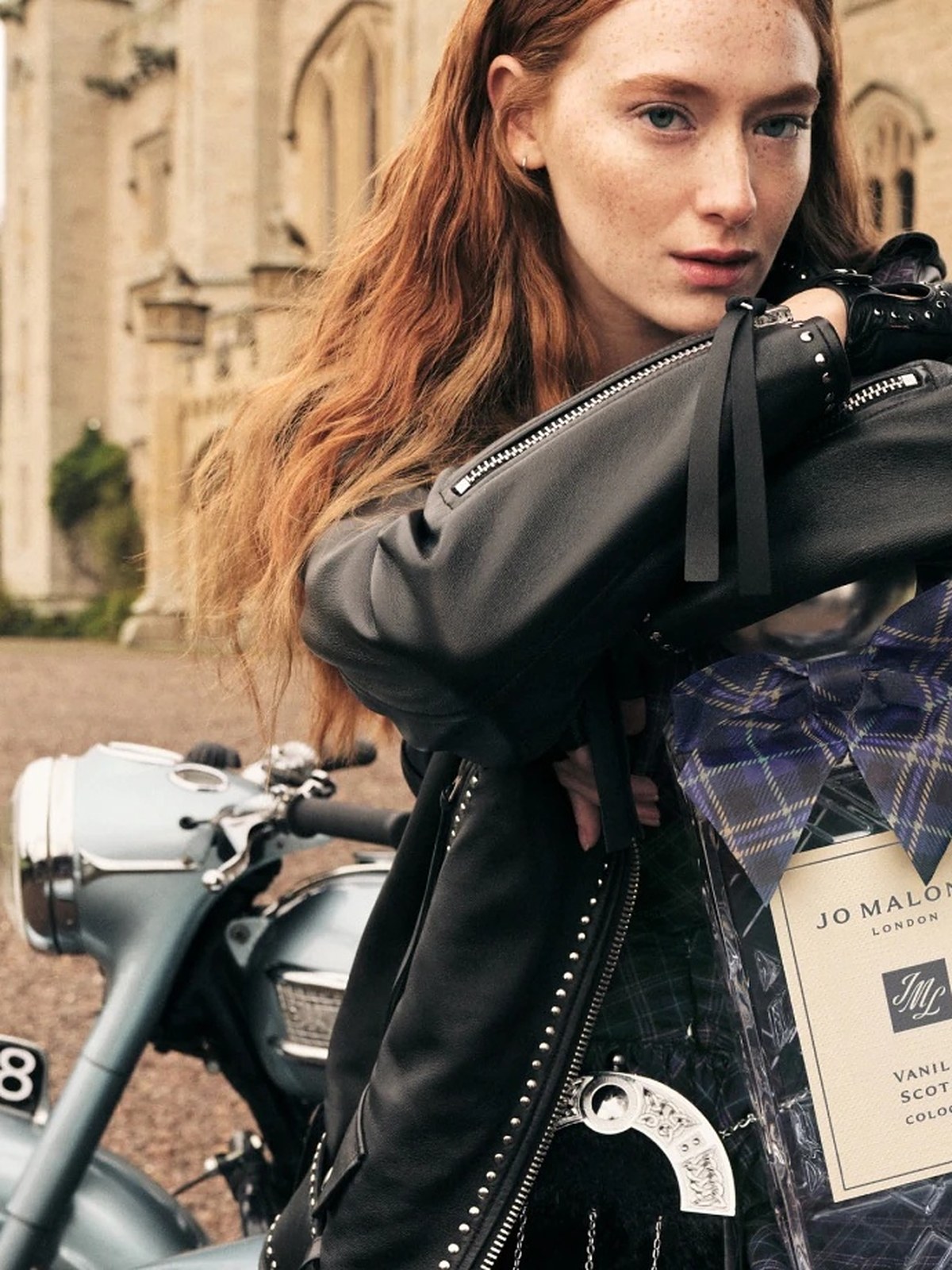 Red haired model in a black leather jacket holding a tartan cologne box by a vintage motorbike