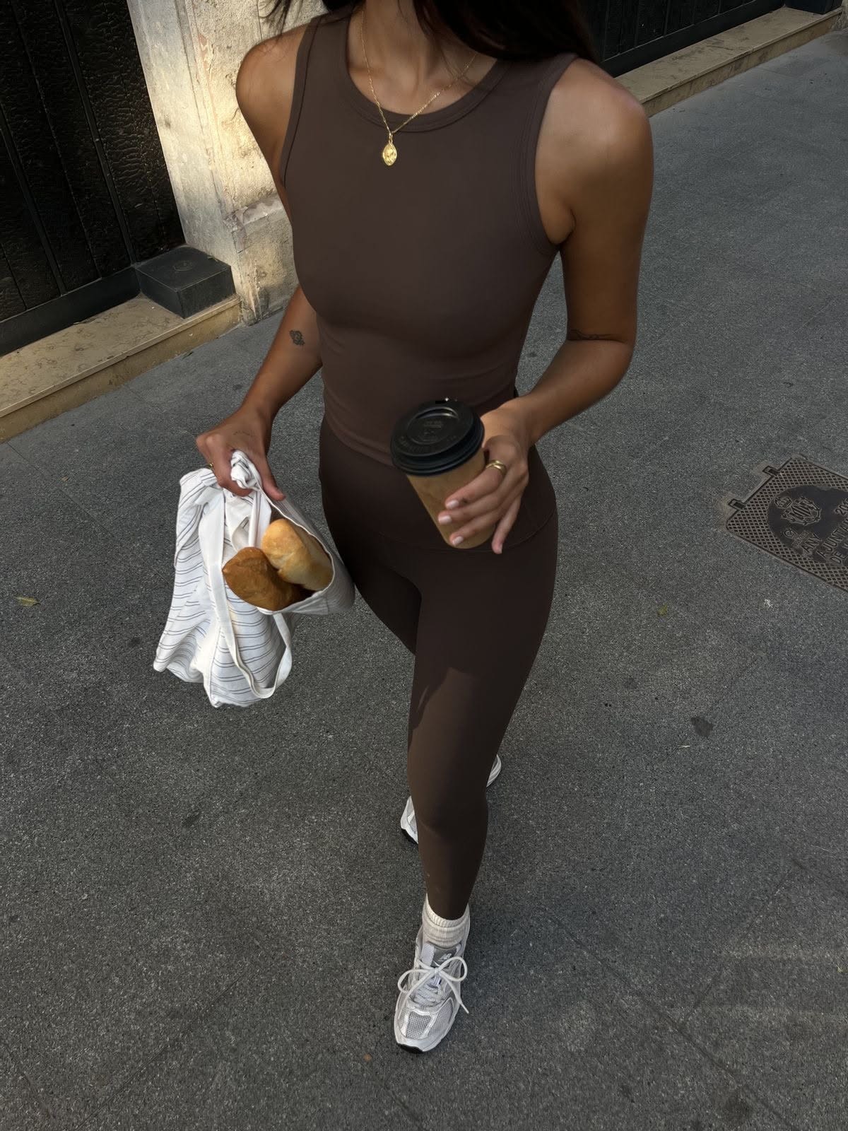 Woman in a brown fitted onesie carrying a coffee and a towel on a city street