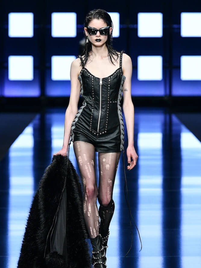 Runway model in a black leather mini look under bright grid lights