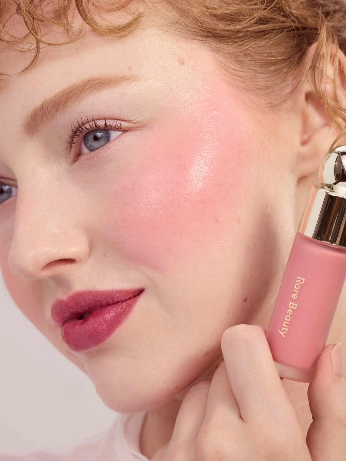 Red-haired model with soft pink liquid blush holding the bottle at her cheek