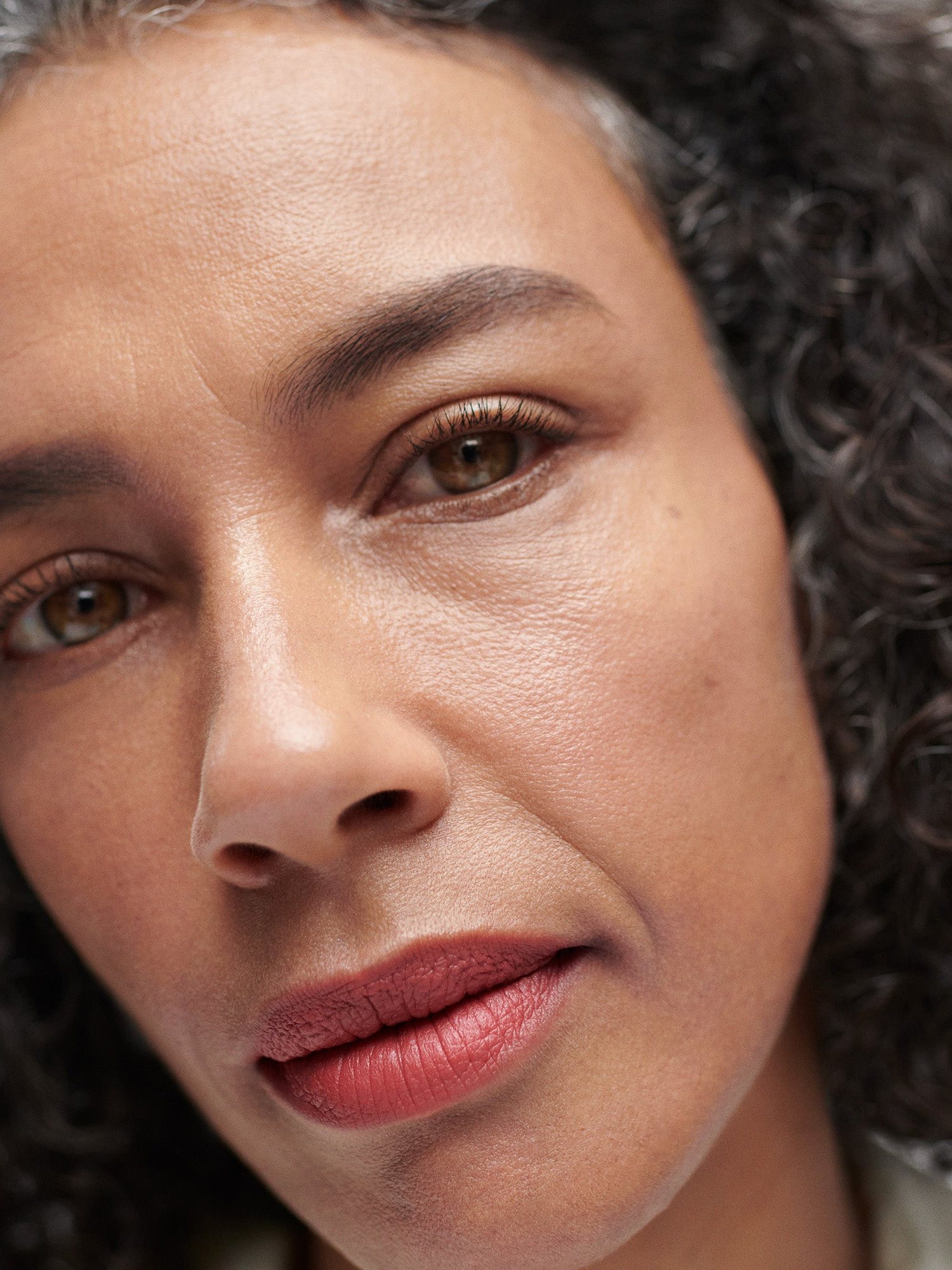 Close up of a woman with curly grey streaked hair wearing a terracotta lipstick