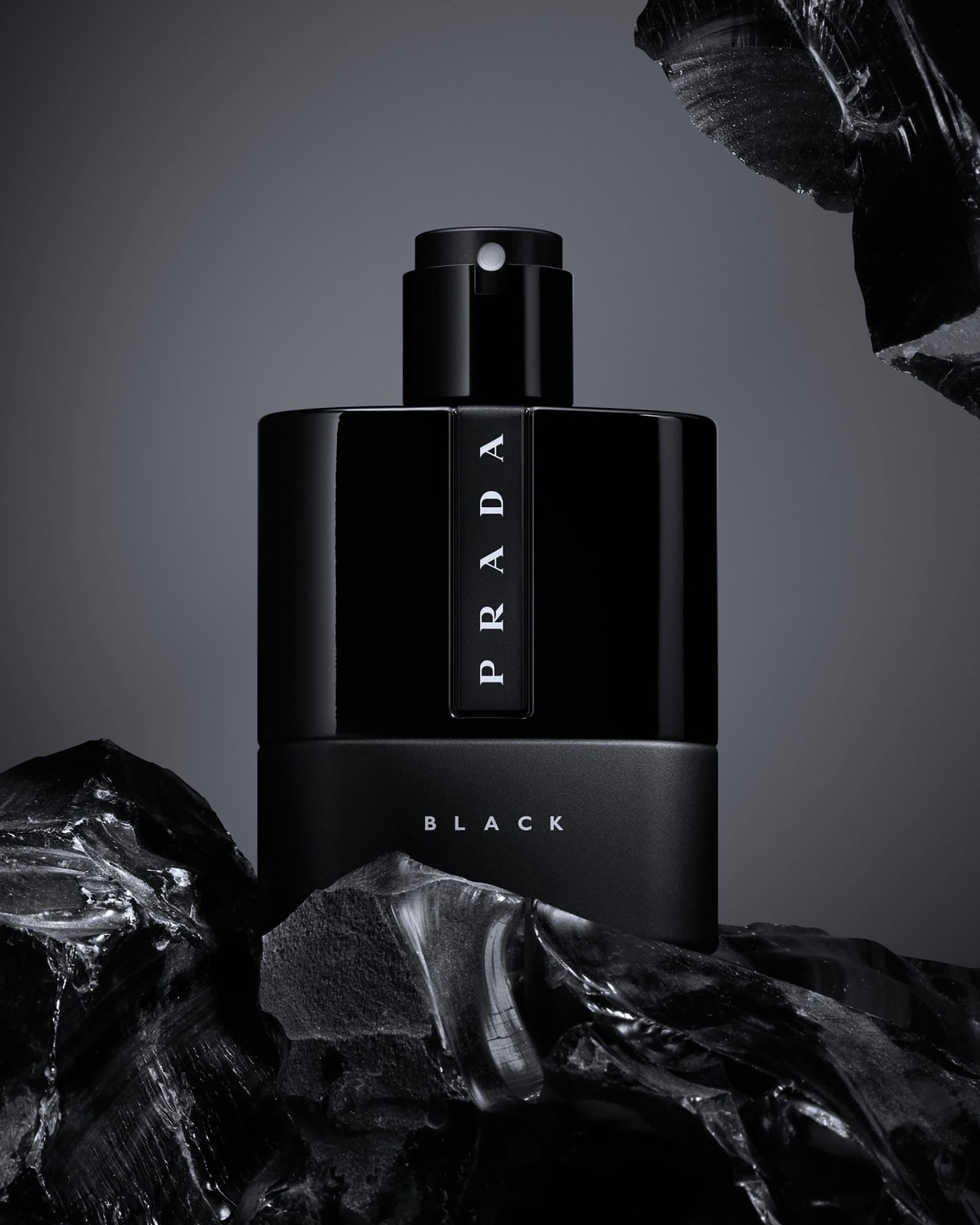 A black Prada Luna Rossa Black bottle photographed against a grey backdrop and shards of black glass